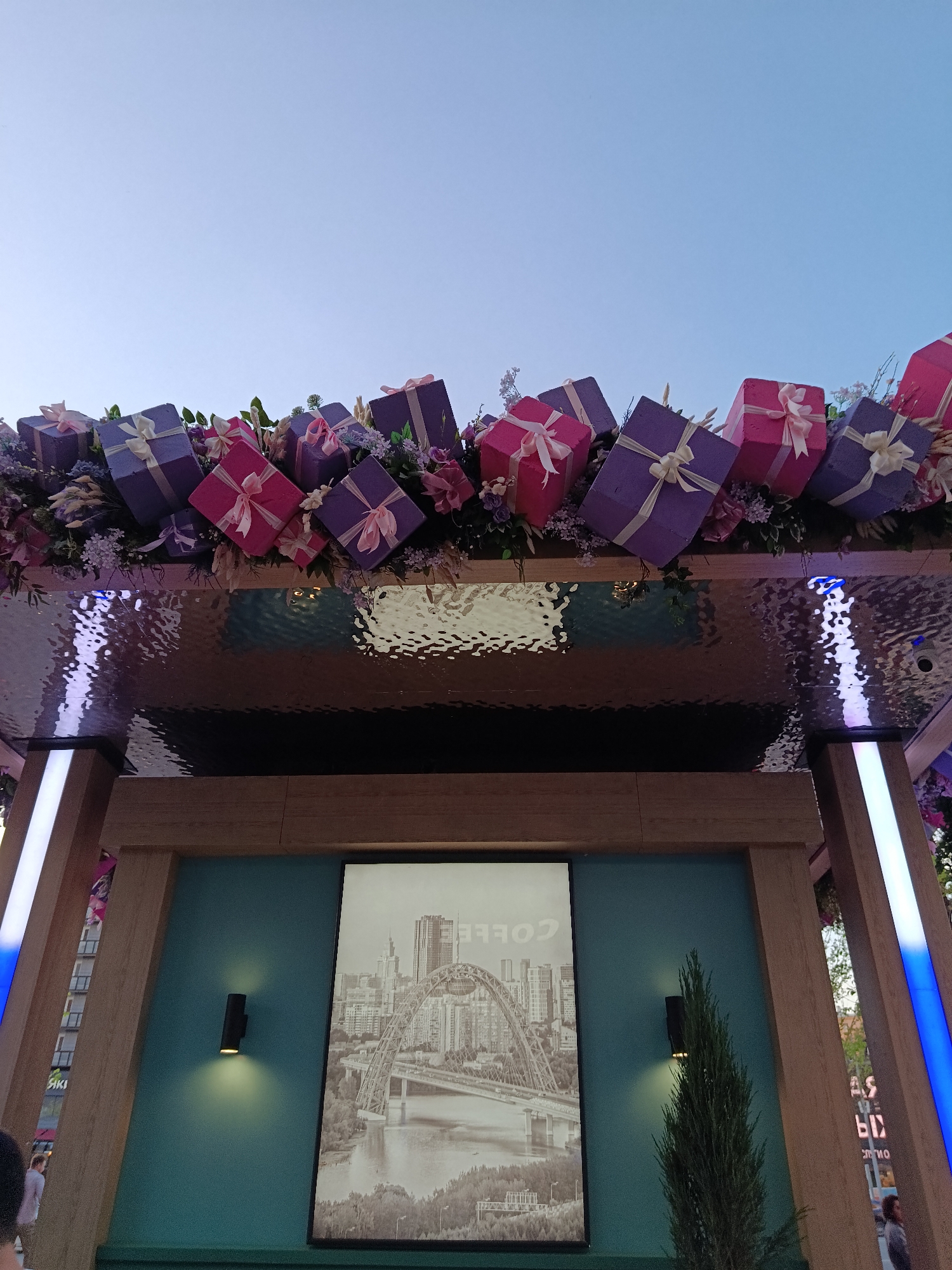 A low-angle shot captures an outdoor structure adorned with decorations, likely a commercial venue. The clear sky in the background suggests either daytime or well-lit conditions. On top of the structure is a garland of colorful gift boxes in shades of pink and purple, accented with flowers. A reflective metallic surface mimics the ripple of water.
Below, the structure features wooden beams framing a teal backdrop. A black-and-white photo depicting a cityscape with a prominent bridge hangs on the teal wall. Small black sconce lights flank the picture. To the right, a coniferous shrub stands, and a blue-lit vertical beam is visible, as is what appears to be advertising in Russian writing.
Based on the bridge and architecture in the black-and-white photo, this appears to be in the city of Moscow, Russia.