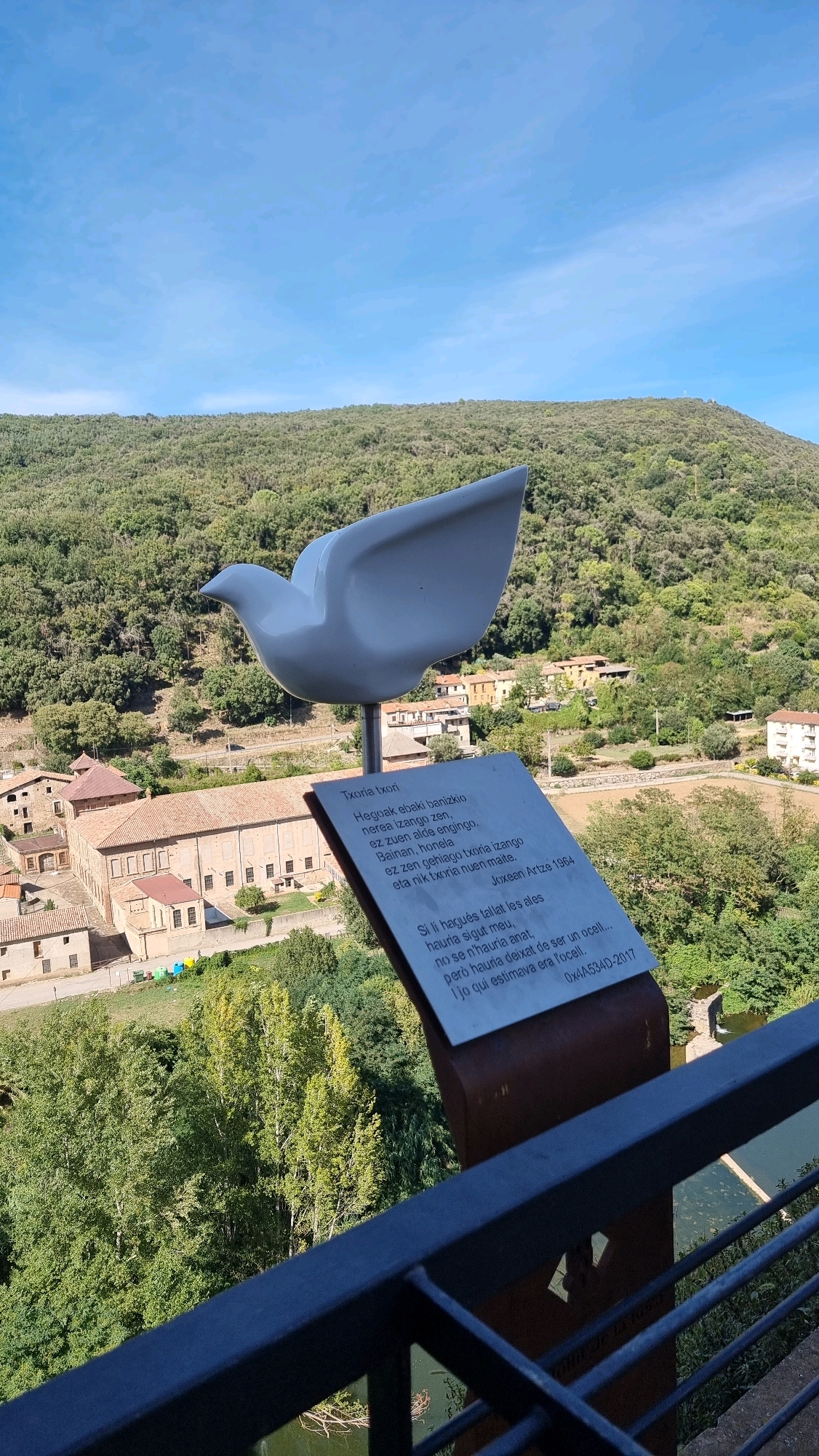 In Castellfollit de la Roca, Spain, a white dove sculpture is positioned overlooking a verdant landscape. The sculpture is mounted on a metal stand inscribed with text in Basque. Behind the sculpture, traditional stone buildings with terracotta roofs are visible, nestled amongst lush green trees. A gently winding river or body of water can be seen in the foreground, bordered by a dark metal railing. The sky above is a clear, bright blue with a few wispy clouds.