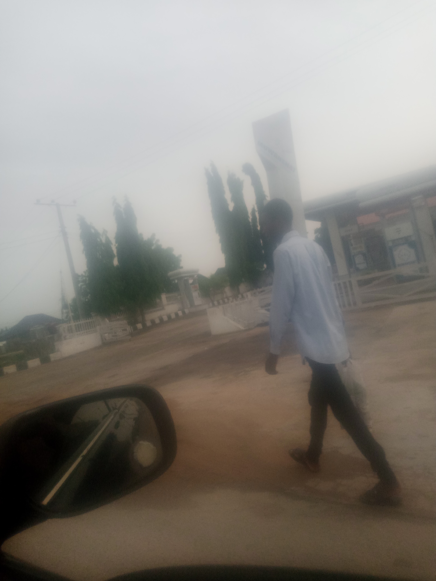 The image captures a pedestrian walking on a dusty street in Jalingo, Nigeria. The sky is heavily overcast, creating a diffused, hazy atmosphere that obscures details of the surroundings. In the foreground, the side mirror of a vehicle is visible, reflecting a portion of the scene. The pedestrian, a young man wearing a light blue collared shirt and dark trousers, walks away from the viewer, carrying a white bag over his right shoulder. He is walking on a dirt road, and behind him, a white-fenced structure with some trees and a lamppost can be discerned through the haze. The overall scene suggests a somewhat arid environment, typical of parts of Jalingo.