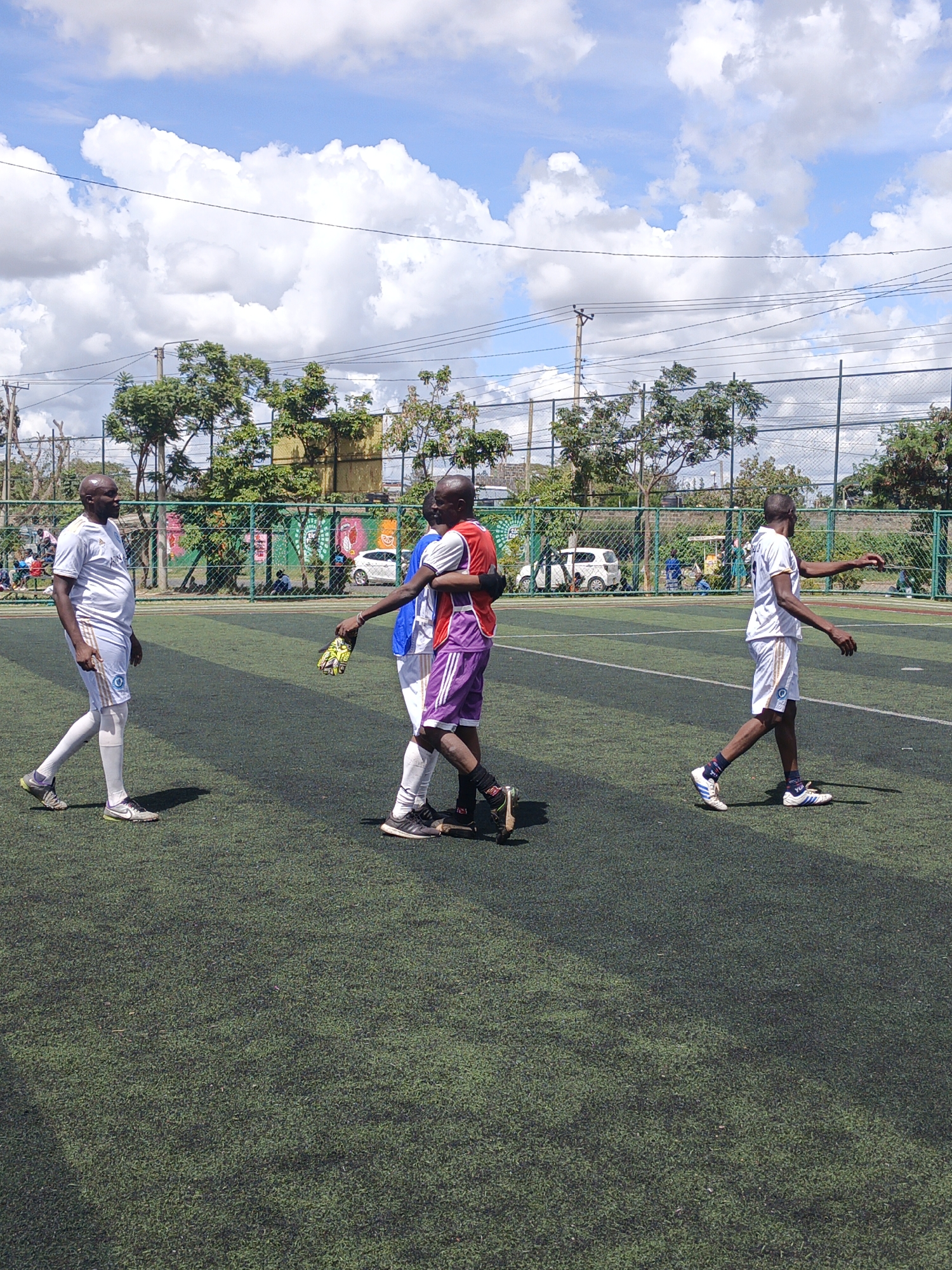The image depicts a soccer field in Nairobi, Kenya. Four male individuals are visible on the green turf, marked with white lines. Two individuals are embracing, while the other two are walking away. All are wearing soccer attire. A chain-link fence surrounds the field, with trees and buildings in the background. A white car is visible behind the fence. The sky is blue with scattered clouds.
