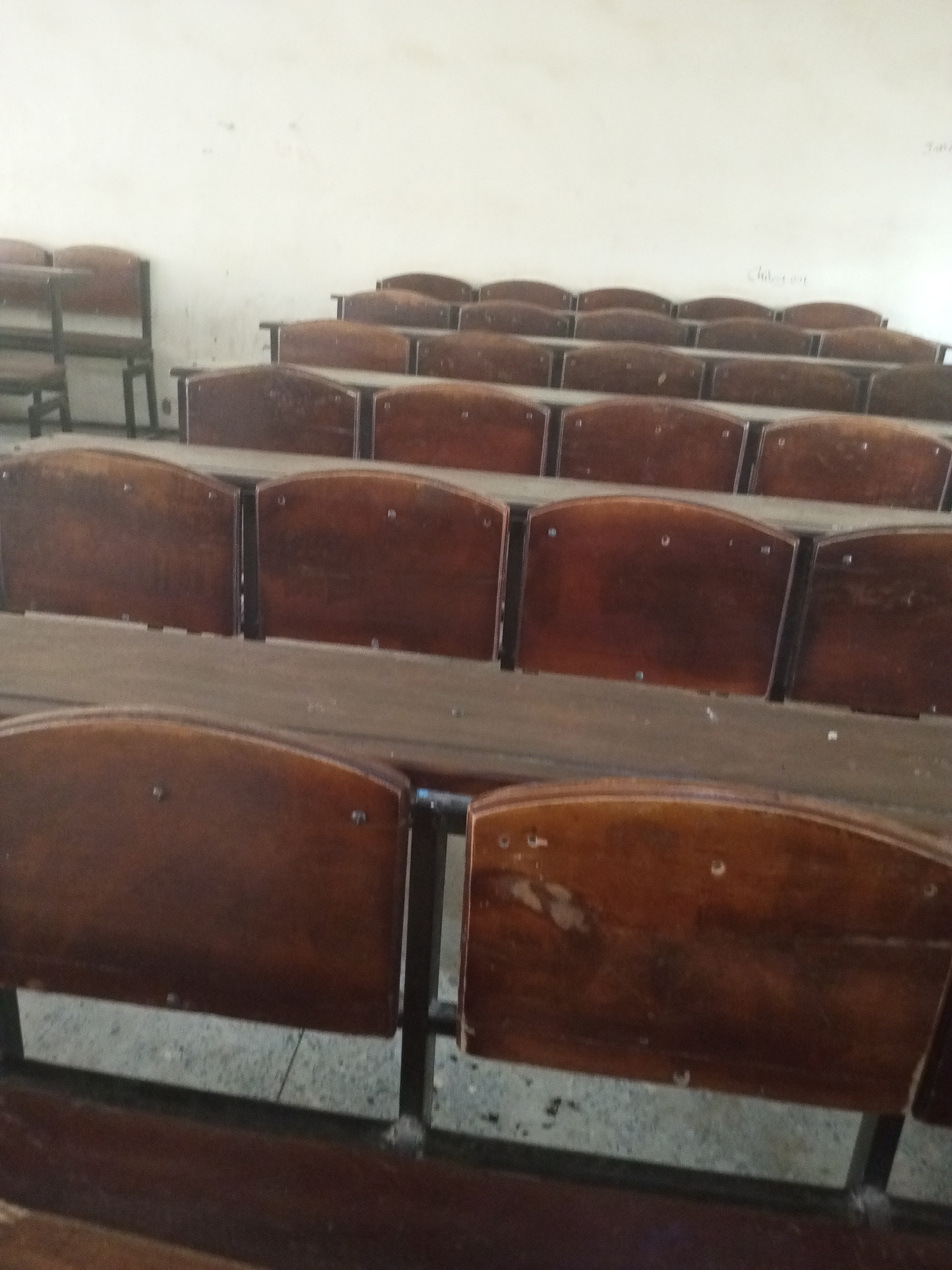 This media file depicts an interior view of an empty lecture hall with tiered seating. The subjects are rows of wooden chairs with dark metal frames. The scene is a deserted classroom, suggesting a break between classes or an unoccupied lecture hall. Notable details include the worn appearance of the wooden seats, indicating age and frequent use. The lighting appears to be artificial, providing a consistent, neutral illumination that doesn't suggest a specific time of day. The background consists of a plain, light-colored wall, possibly painted plaster, with some faint, illegible writing or markings. There are no people present, and therefore no activity or emotions are visible. The setting appears to be a typical academic environment, potentially a university or college lecture theater. The provided location context of Jalingo, Nigeria, suggests this is a lecture hall within an educational institution in that region. No discernible text is visible in the image.