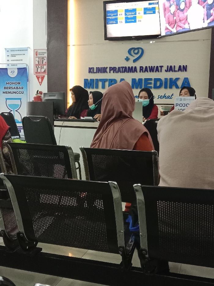 The image depicts the waiting area of Klinik Pratama Rawat Jalan Mitra Medika, located in Gladaksari, Indonesia. Six individuals are visible: three staff members behind a counter and three individuals seated in the waiting area.
The staff members, all women wearing hijabs, are positioned behind a dark counter on the right side of the frame. The leftmost staff member is partially obscured, facing a computer monitor. The second staff member from the left wears a dark hijab and a light blue medical mask, looking at a monitor. The third staff member, also in a dark hijab and a light blue medical mask, looks to the right. The fourth staff member, furthest right, wears a dark hijab and holds a sign that partially reads "POJO".
In the foreground and midground, three individuals are seated in black perforated metal waiting room chairs with chrome accents. The person in the immediate foreground is seen from the back, wearing a light beige or brown garment. Directly in front of the counter, a person is seated with their back fully visible, wearing a reddish-brown hijab and matching top. A third person's red top is partially visible to the far left, obscured by the seating.
Above the counter, a large blue sign displays "KLINIK PRATAMA RAWAT JALAN MITRA MEDIKA" with a blue heart-and-plus logo. Above this, a digital queue display screen shows various numbers (e.g., 44, 6, 16, 36) under categories like "POLI UMUM" and "POLI GIGI." To the far left, a blue and white banner reads "MOHON BERSABAR MENUNGGU" (Please Be Patient Waiting) with an hourglass icon, next to a red fire extinguisher instruction sign and a small fire extinguisher. The floor is tiled, and the walls are predominantly white with some vertical accent lighting.