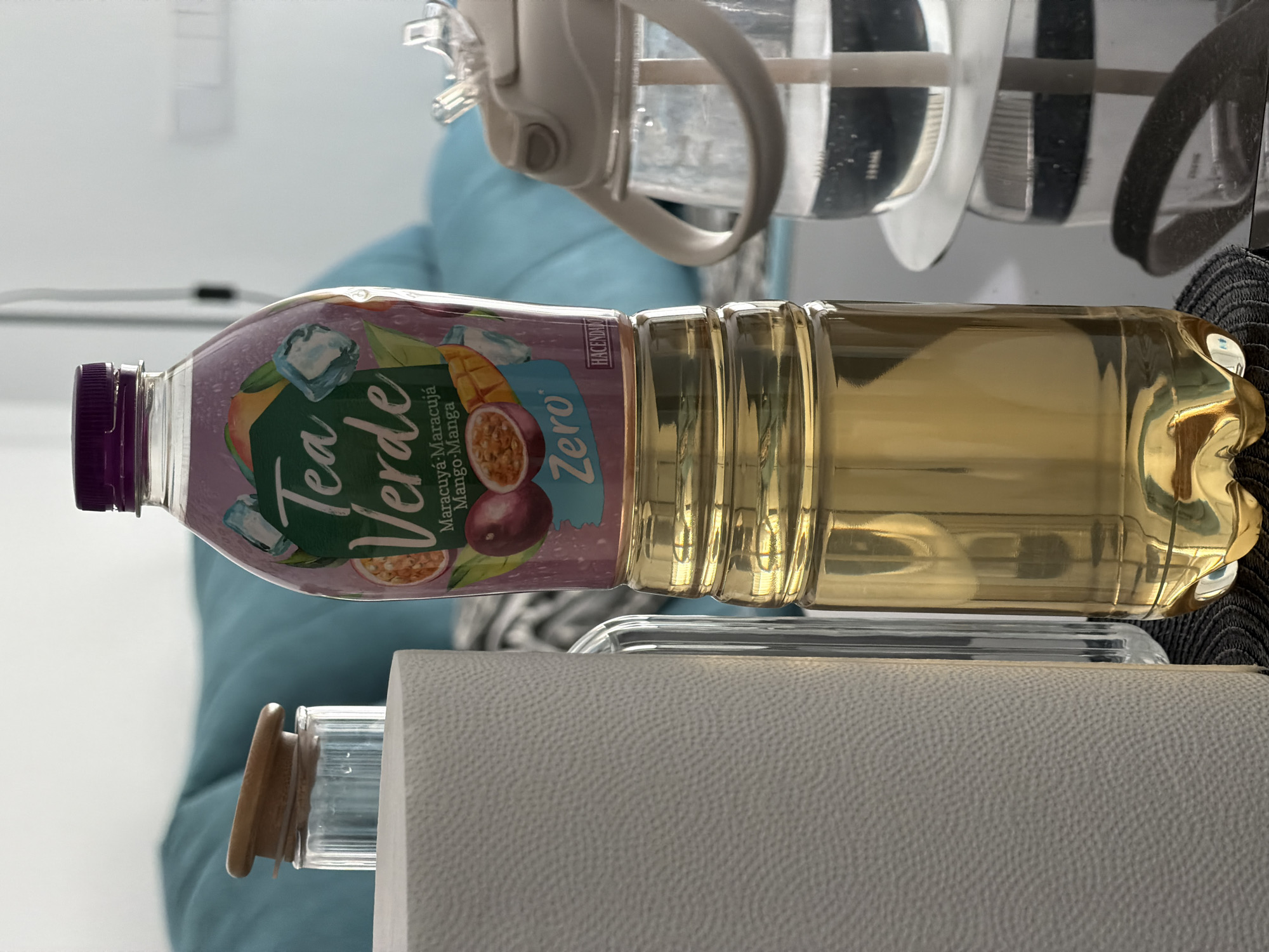 The image shows a close-up of several objects on a dark, reflective surface, likely a table. In the foreground, on the left, is a white, textured roll of paper, possibly paper towels. To its right, a clear glass container with a wooden lid is visible, containing a small amount of liquid and condensation. Directly behind these, a tall, clear glass bottle stands partially obscured.
The central and most prominent object is a plastic bottle of "Tea Verde" with a purple cap. The bottle's label is colorful, featuring illustrations of passion fruit, mango, and ice cubes, with the text "Maracuyá-Maracujá Mango-Manga" and "Zero" prominently displayed. The lower portion of this bottle contains a golden-yellow liquid.
To the right of the "Tea Verde" bottle, a clear plastic water bottle with a light-colored lid and straw is visible, containing a dark liquid. The background is out of focus but suggests an indoor setting with a blurred blue sofa.
The image was captured in Finestrat, Spain. The lighting is bright, with reflections indicating an overhead light source. There are no people present in the image. The scene appears to be a still life arrangement of beverages and household items.