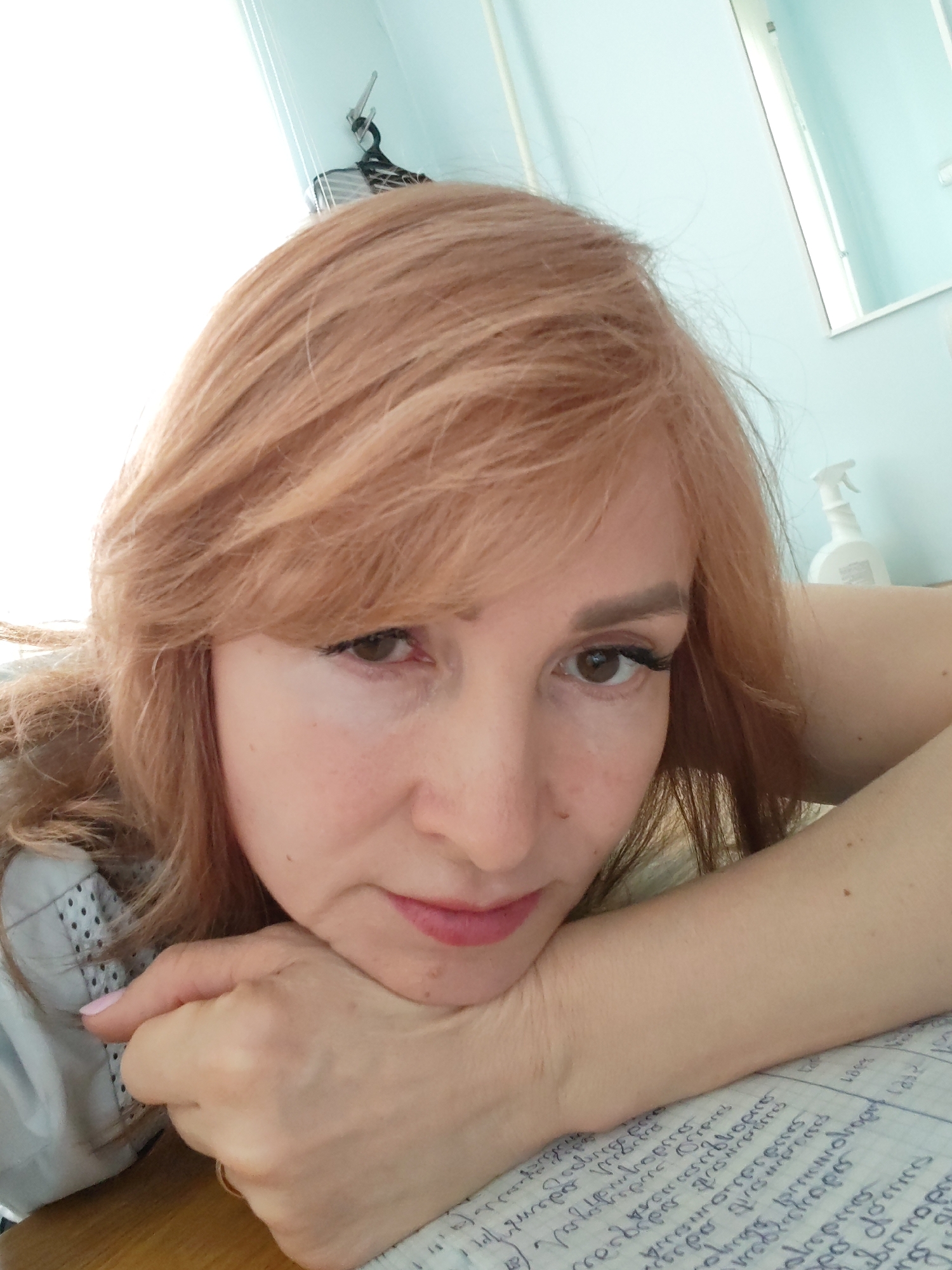The image depicts an indoor scene in the city of Chaikovsky, Russia, featuring an adult female subject. The woman, with light auburn hair styled with bangs, is centrally positioned, looking directly at the viewer. Her dark brown eyes are visible, enhanced by eyeliner and false eyelashes. Her eyebrows are shaped and filled. Her lips are closed and show a light pink hue. She rests her chin on her clasped hands, which are placed upon a wooden surface. Her left hand displays light pink nail polish, and a gold-colored ring is present on the ring finger of her right hand. She is wearing a light-colored garment with a visible dotted texture on the left sleeve. Her hands and forearms rest over a grid-ruled sheet of paper containing handwritten blue text, which appears to be in Cyrillic script. The background consists of a light blue wall. In the upper right quadrant, a white-framed mirror is visible, reflecting the light blue wall. A white spray bottle is positioned on a surface below the mirror. A bright, indistinct light source, possibly a window, occupies the upper left background. A dark, possibly striped object hangs from a hook against the light blue wall behind the subject's head.