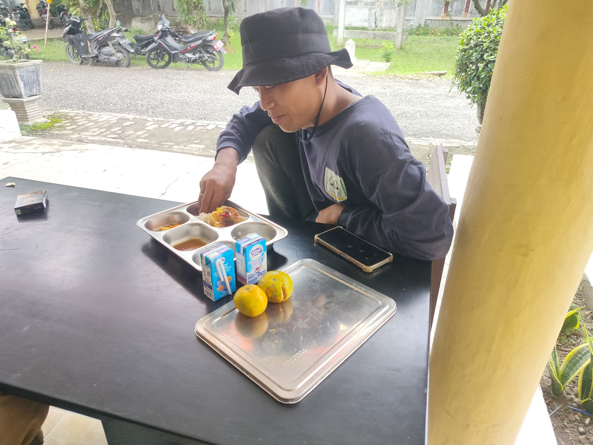 A medium shot shows a man wearing a black bucket hat and a dark blue long-sleeved shirt, sitting on a bench in front of a black table. He is leaning forward and using his right hand to pick up food from a metal tray with multiple compartments. The tray contains rice, a fried item, and some sauce. Two small cartons of milk and two oranges, one peeled, are on the table. A cell phone is also on the table. In the background, several motorcycles are parked on a gravel path. A potted plant is to the left of the motorcycles, and a yellow pillar is to the right of the frame. The scene appears to be outdoors, possibly in Lawebekung, Indonesia, with some greenery and a building structure visible in the distance.
