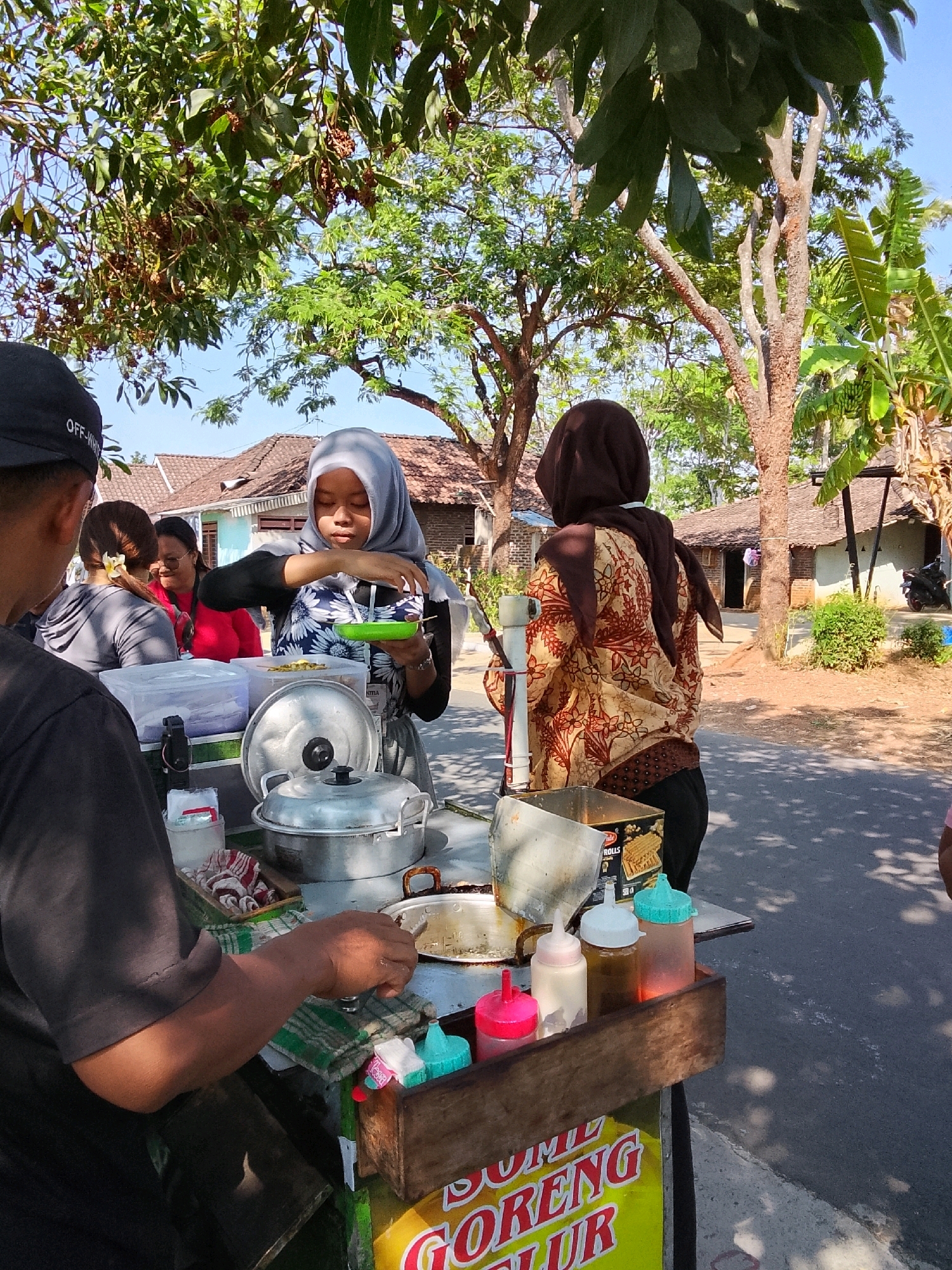 The image depicts an outdoor scene in Kaliwungu, Indonesia, likely a roadside stall. In the foreground, a person wearing a black t-shirt and a black baseball cap faces away from the camera, their arm extended towards a food cart. The cart itself is laden with various food items and containers. A woman wearing a gray hijab and a floral-patterned shirt is actively serving food, holding a green bowl and a utensil. Behind her, another woman in a brown headscarf and a patterned jacket also appears to be involved in the stall's operations. Several other individuals are visible in the background, along with residential buildings and lush green trees. The overall impression is of a bustling, informal marketplace.