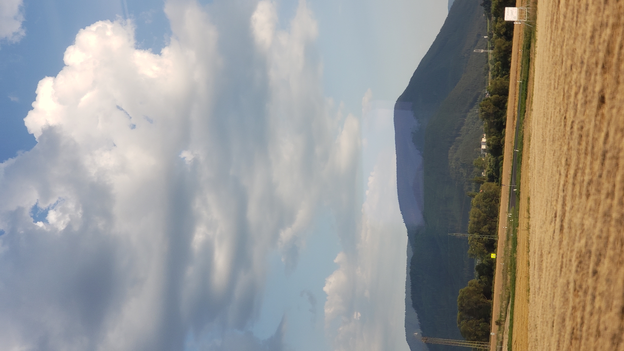The image shows a rural landscape with a field of dry, golden grass in the foreground. In the middle ground, there is a line of trees and a road. In the background, there are rolling hills covered in dark green forests. The sky is partly cloudy with large white clouds against a blue sky. There are no people or animals visible in the image. The scene is peaceful and appears to be taken during daylight. There is no specific information about the location in the city of Beňadiková, Slovakia, within the image itself.