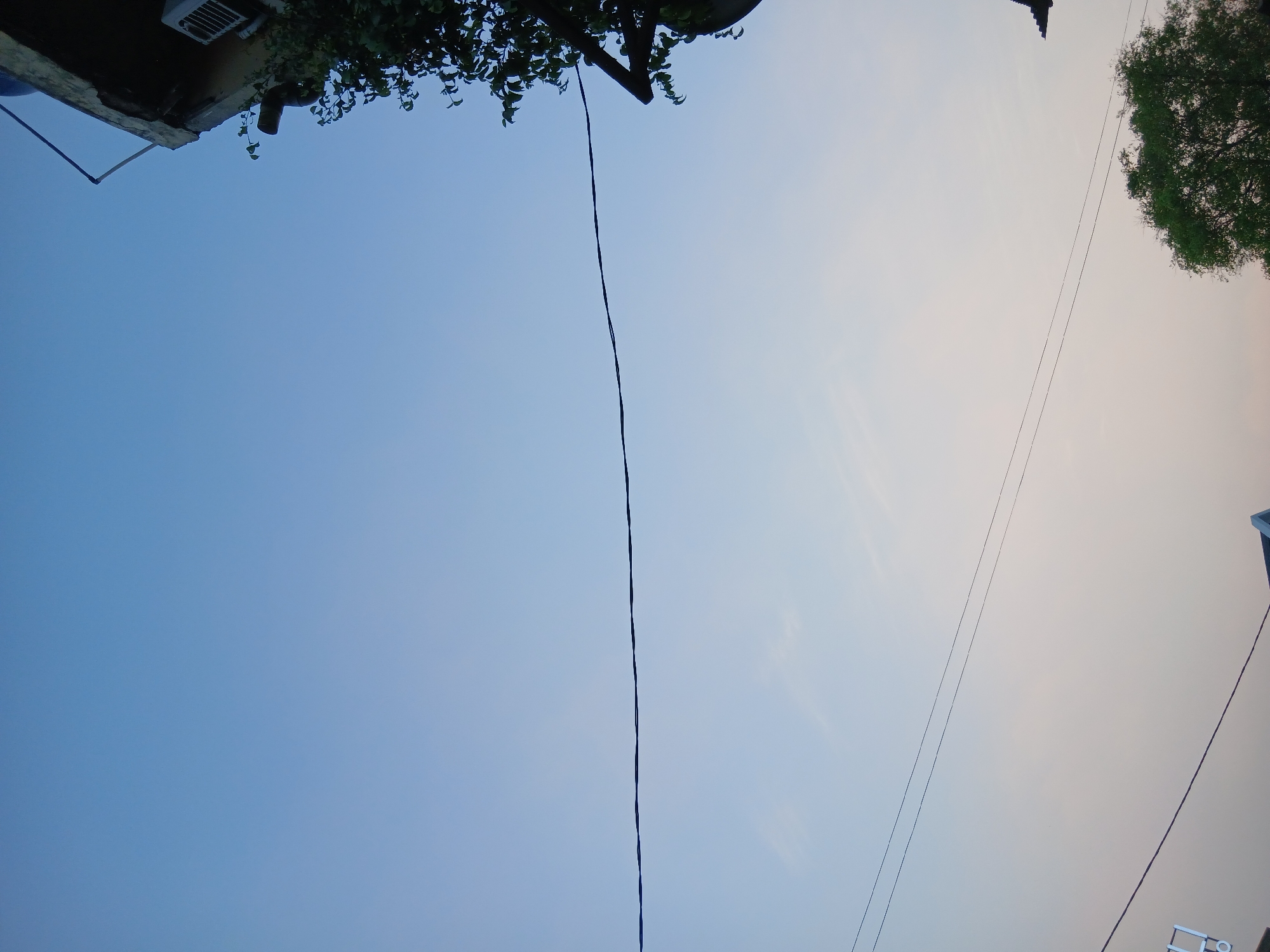 The image depicts a clear sky with a gradient from a lighter blue at the bottom to a darker blue at the top. A single black wire stretches horizontally across the middle of the frame. In the upper right corner, a portion of a building is visible, with a weathered concrete edge and an air conditioning unit attached to its side. Lush green foliage from a tree hangs over the building's edge. In the lower right corner, the canopy of a mature tree with dense green leaves is prominent. Two more thin black wires are visible running parallel to each other in the lower half of the image, and a faint outline of a roofline can be seen at the very bottom. The scene appears to be an outdoor view, possibly from a residential area, with elements suggesting an urban or suburban environment in South Tangerang, Indonesia. There are no people or specific actions being performed.
