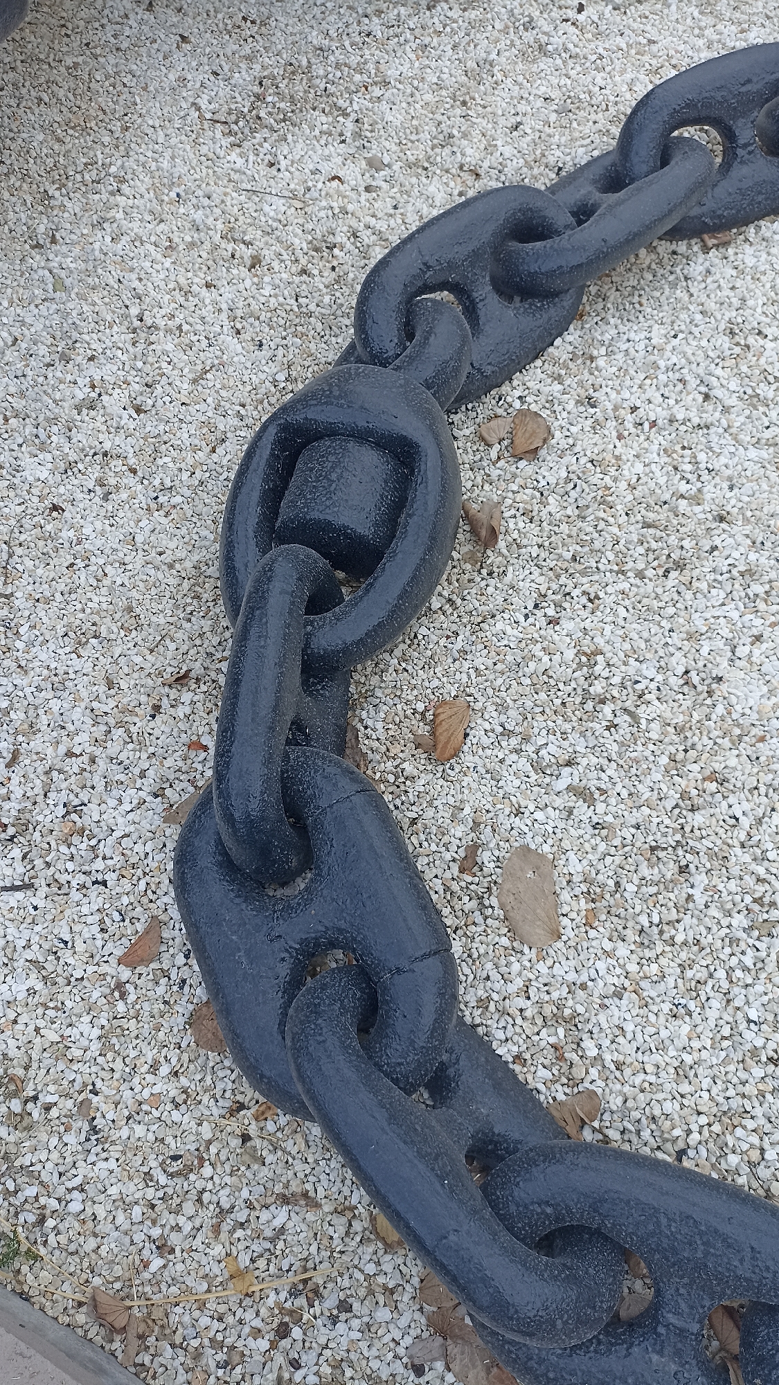 A massive, dark grey or black chain lies prominently across a light-colored gravel surface in this high-angle, close-up photograph. The chain's links are exceptionally thick and robust, suggesting it is a heavy-duty anchor chain or part of a large mooring system, commonly found in maritime environments. Many of the links feature a central stud or bar, a design element typical for anchor chains to enhance their strength and prevent twisting. The chain's surface appears textured, likely from paint, giving it a somewhat matte finish.
The ground beneath the chain is covered with small, irregular, off-white or light grey pebbles. Scattered amongst the gravel are several dry, brown leaves and a few small twigs, indicating an outdoor setting. The diffused, even lighting suggests it is daytime, possibly under overcast conditions, as no harsh shadows are cast.
No human activity or event is taking place; the chain appears to be a stationary display or a permanent fixture. The absence of people and visible text allows the focus to remain entirely on the industrial scale of the chain and its immediate environment. The location context of Yenişehir, Turkey, a district known for its port, aligns well with the presence of such a large maritime chain.