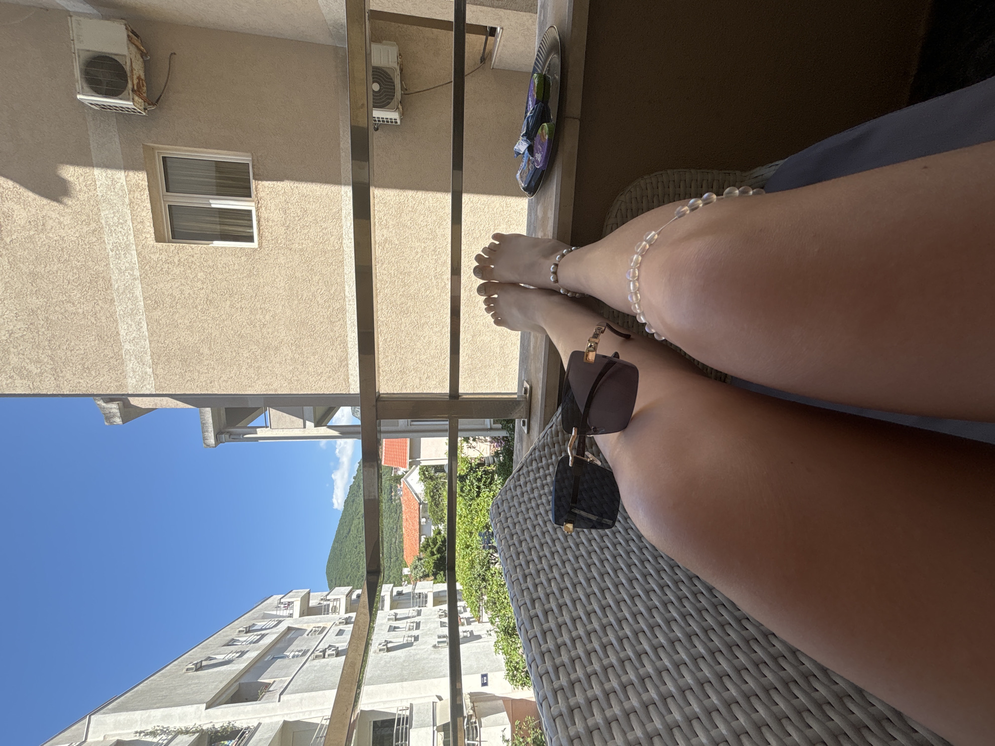 The image presents a first-person perspective from a balcony in Budva, Montenegro, on a clear, sunny day. In the immediate foreground, a person's bare, tanned legs are extended, with the feet resting on a narrow ledge of the balcony. One ankle displays a light-colored, beaded anklet, possibly made of pearls. Dark-lensed sunglasses with gold-colored accents are placed on the right thigh. The legs are positioned over a light grey, woven patio chair.
To the right of the feet on the balcony ledge, a silver, oval-shaped tray holds several individually wrapped, colorful items, likely chocolates or candies, next to a pair of multi-colored flip-flops. A horizontal metal railing extends across the mid-ground, separating the balcony from the view beyond.
The surrounding environment consists of residential buildings and natural landscape. To the left, a light-colored, multi-story apartment building with multiple balconies and external air conditioning units is visible. To the right, a beige, textured building wall with a rectangular window and an older, rusted air conditioning unit is partially illuminated by direct sunlight, casting a sharp vertical shadow across its surface. In the mid-distance, additional residential structures with red-tiled roofs are interspersed among dense green trees and foliage. The background features a large, prominent green mountain covered in vegetation, beneath a vast, clear blue sky with a few small white clouds.