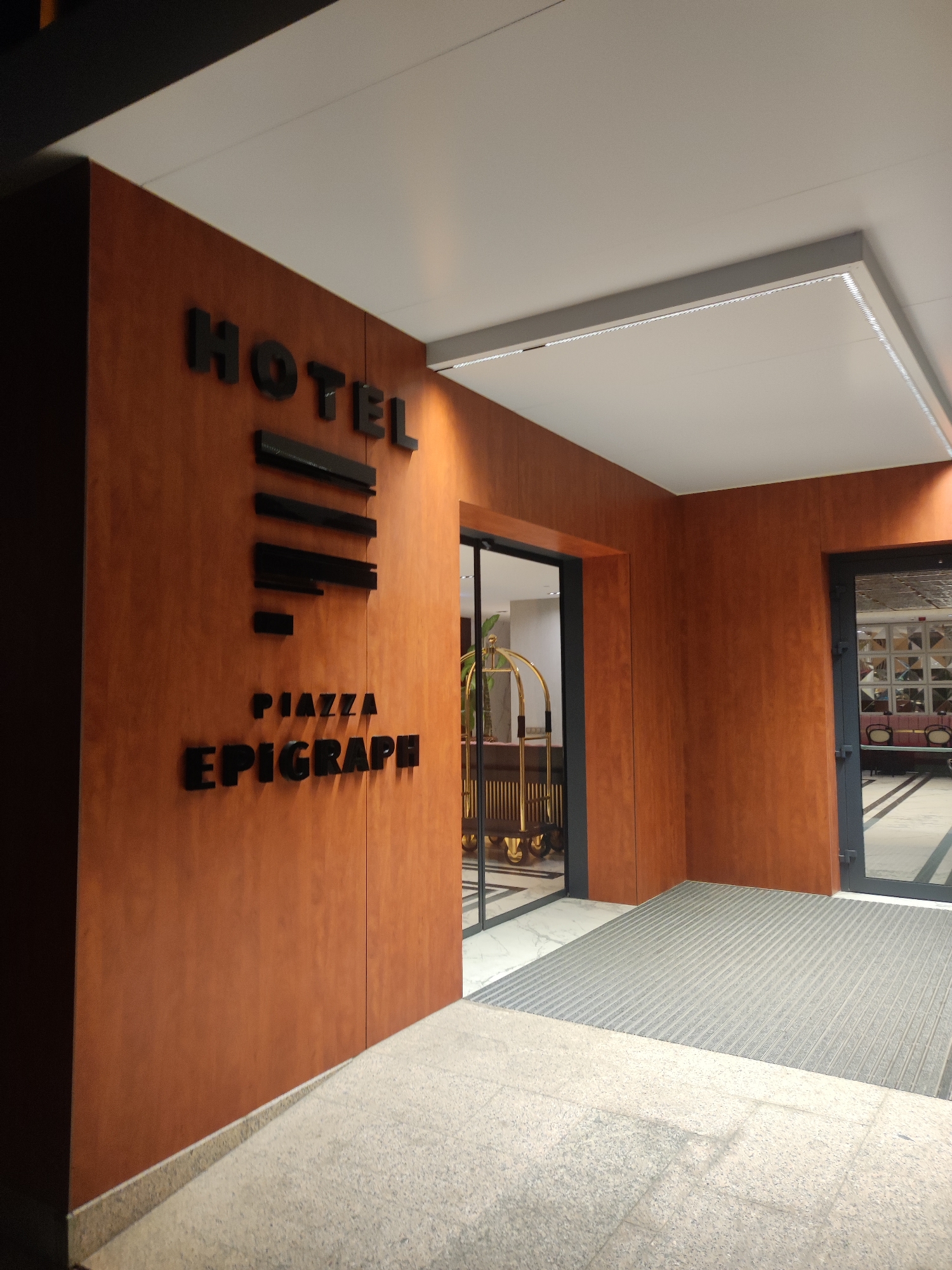 The image shows the exterior of a hotel named "HOTEL PIAZZA EPIGRAPH" in Batumi, Georgia. The hotel has a modern architectural design with a facade covered in wood-like panels. The entrance is marked by a prominent sign with black 3D letters spelling out the hotel's name. To the left of the entrance, a glass door leads into the hotel lobby, where a brass luggage cart is visible. To the right, another glass door opens into a seating area with upholstered benches and tables. The floor is paved with granite tiles and a grey ribbed mat is placed at the entrance. The ceiling features embedded LED lighting, creating a warm and inviting ambiance. The overall scene suggests a well-maintained and stylish establishment.
