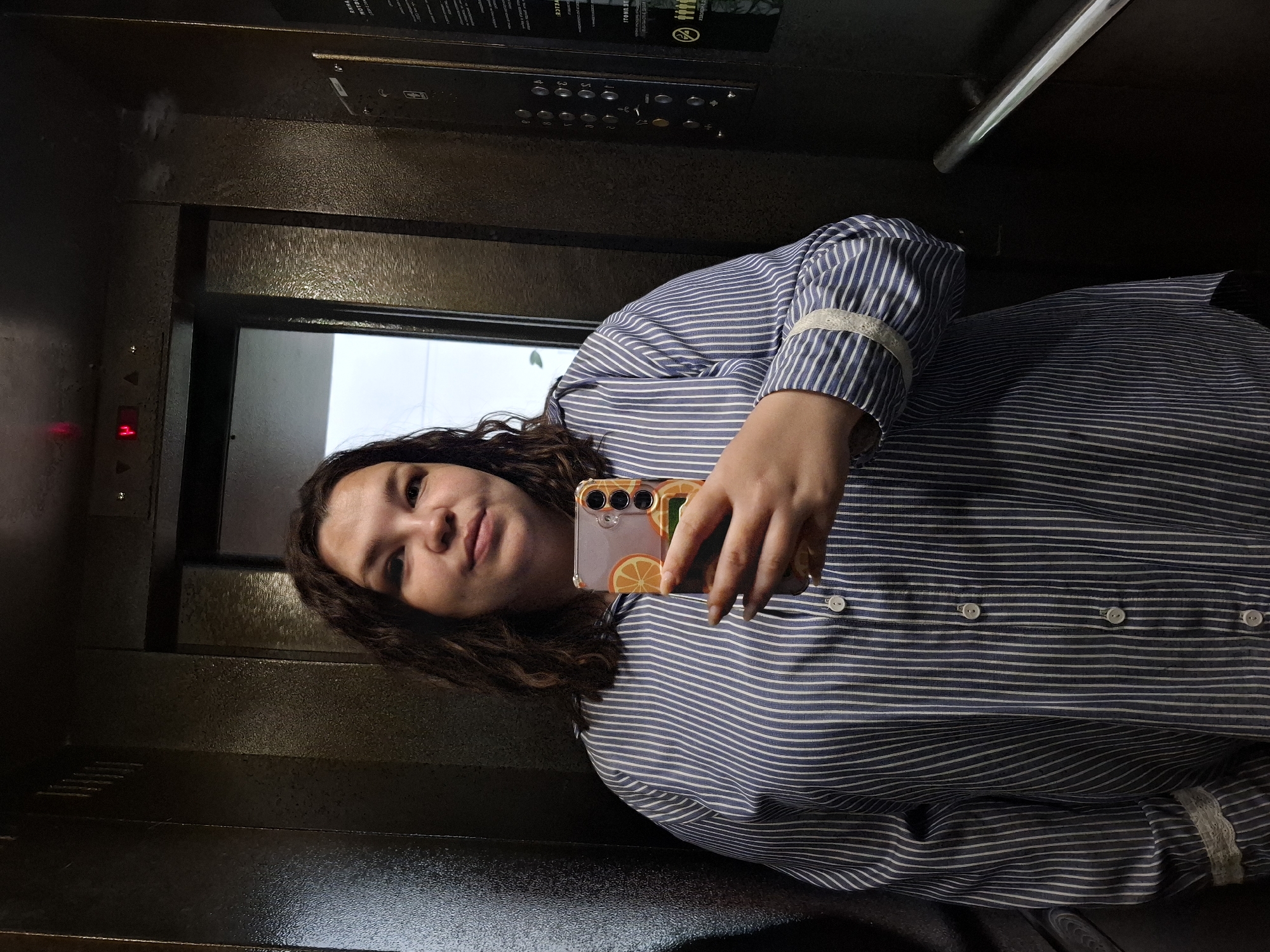 A woman with curly brown hair and wearing a blue and white striped shirt is taking a selfie in an elevator. The elevator has a panel with buttons, a light, and a door. The location is Glazov, Russia, however, cannot be verified from the image.