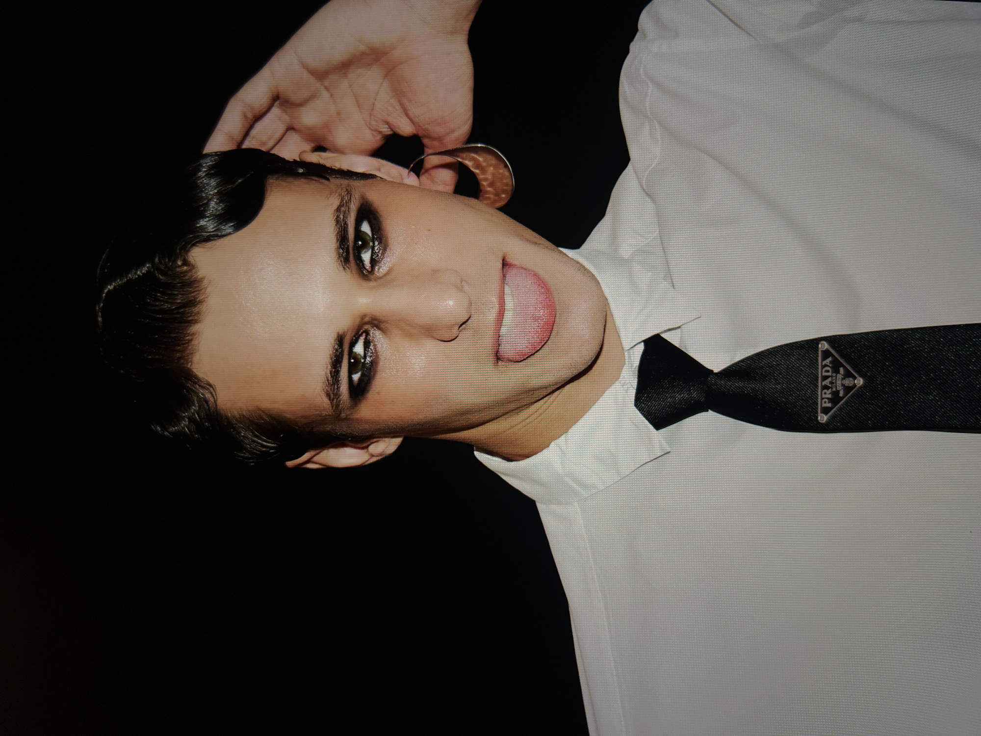 A young person with dark, slicked-back hair and heavy eye makeup sticks their tongue out, holding their right hand to their ear. They are wearing a white collared shirt and a black tie with a silver Prada logo. The background is solid black.