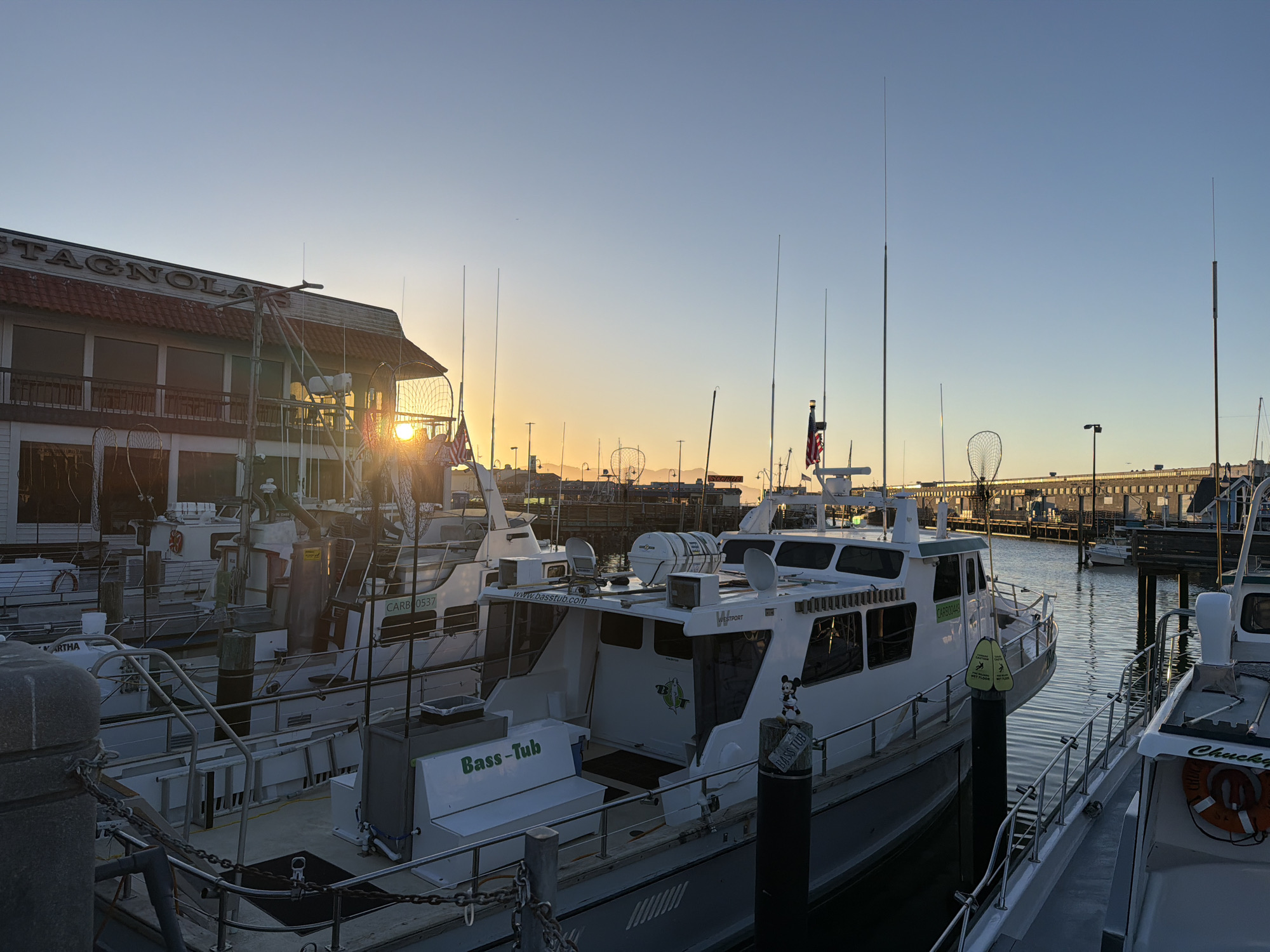 The image depicts a view of a harbor at sunset, likely in San Francisco, California. In the foreground, several white fishing boats are docked side-by-side. The boat on the far right has "Chucky" written on its side and a life preserver with an orange ring. The boat to its left, labeled "Bass-Tub," has a bench with the same name inscribed on it. A small Mickey Mouse figurine is perched on a post next to this boat. The boats have numerous antennae and fishing nets are visible on some.
In the background, a building with the partially visible sign "STAGNOLA'S" is present, suggesting a restaurant or establishment. The setting sun casts a warm glow across the sky, creating a serene atmosphere. The water of the harbor reflects the colors of the sky and the boats. Distant structures and lights are visible along the waterfront, indicating an urban environment. The overall scene is one of a working harbor at the end of the day.