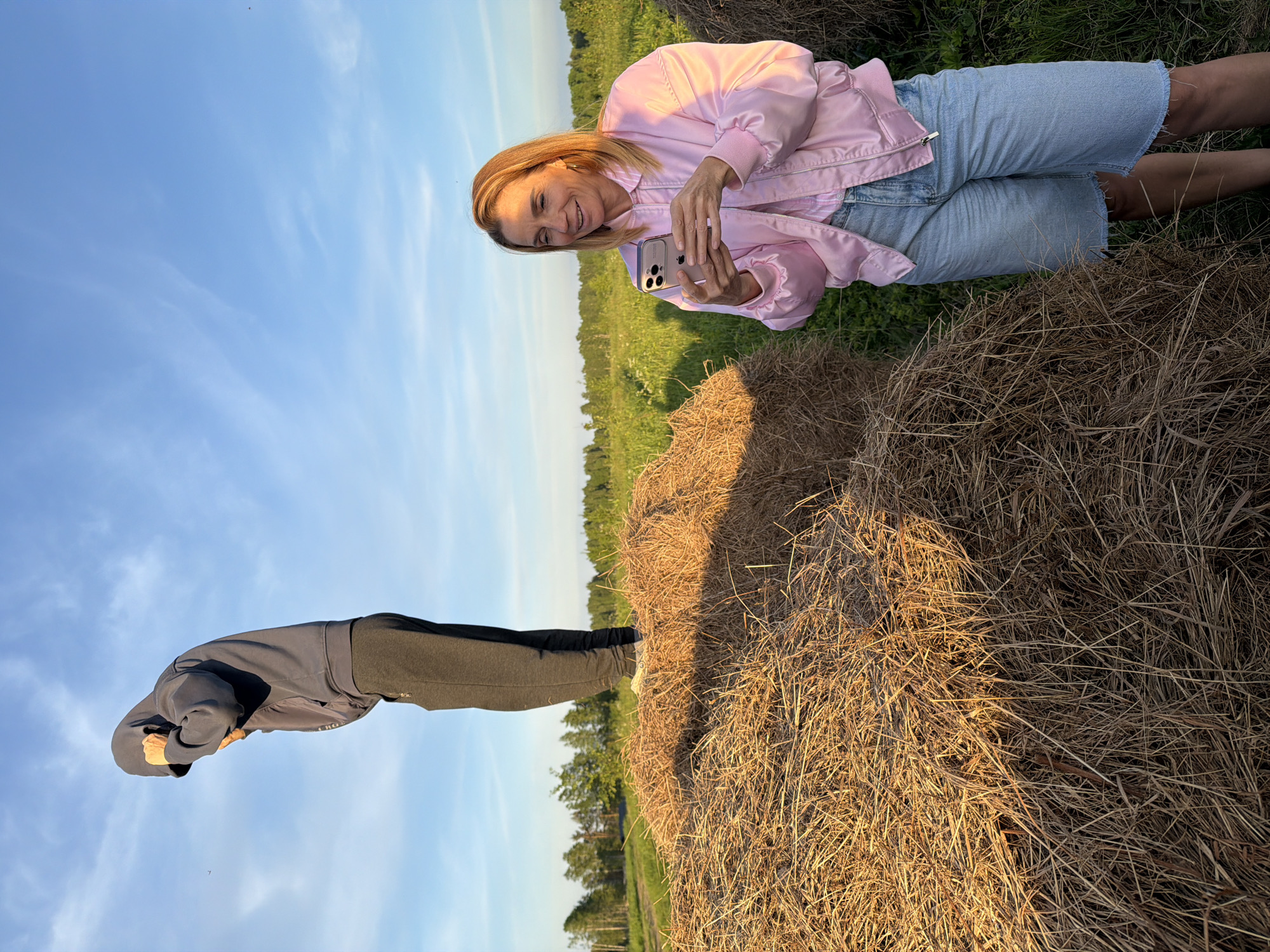 This image depicts an outdoor rural scene during the golden hour, likely late afternoon or early evening, characterized by warm, directional sunlight. The setting is a vast green field with a distant treeline, under a bright blue sky dotted with wispy clouds. Several large, rustic hay bales are prominent in the foreground and midground.
Two individuals are present in the frame. One woman, with blonde hair, is wearing a pink bomber jacket over a light-colored top and denim shorts. She is smiling and looking down at a smartphone, which she holds with both hands, seemingly engaged in taking a photo or video, or viewing content. Her expression conveys happiness and relaxation. The other individual, seen from behind, is wearing a dark grey or black hooded sweatshirt and dark trousers or leggings. This person is in a bent-over posture, with their head lowered, possibly stretching or looking at something on the ground near a hay bale.
The scene captures a moment of leisure in the countryside, likely in Vaskelovo, Russia, given the location context. The hay bales suggest an agricultural area. No visible text is discernible on clothing or objects in the image.
