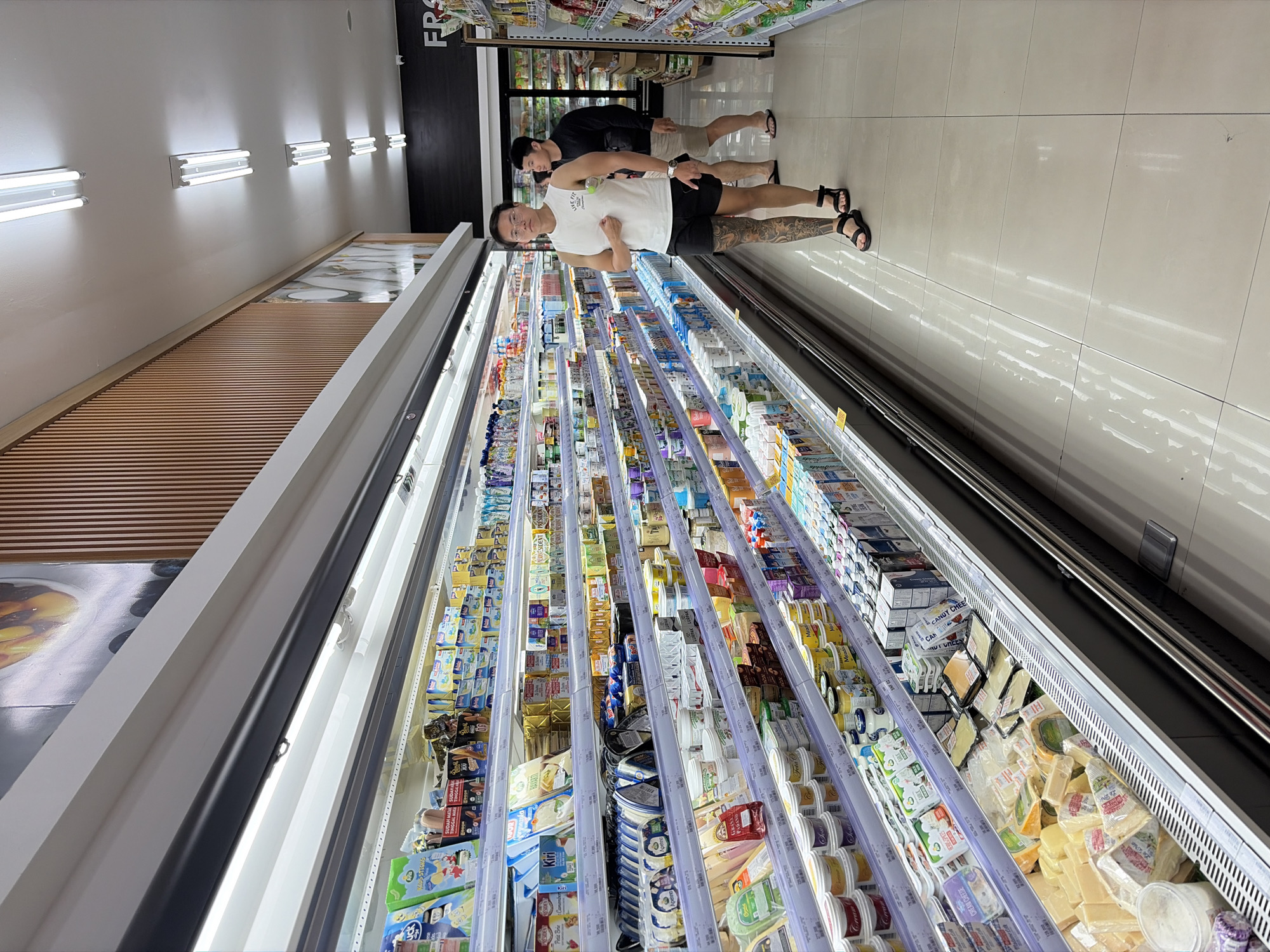 Two men are in a brightly lit supermarket aisle. The aisle is lined with refrigerated display cases filled with dairy products, including various types of cheese, yogurt, and butter. The man in the foreground, wearing a white tank top and black shorts, is walking away from the camera and towards the display cases. He has a tattoo on his left leg. He is wearing sandals. The second man, further down the aisle and behind the first, is wearing a black long-sleeved shirt and khaki shorts and is also walking away from the camera. The supermarket has white tiled floors and fluorescent lighting fixtures on the ceiling. A sign with the word "FRESH" is visible on a black background on the right side of the image. The setting is a supermarket in North Kuta, Indonesia.