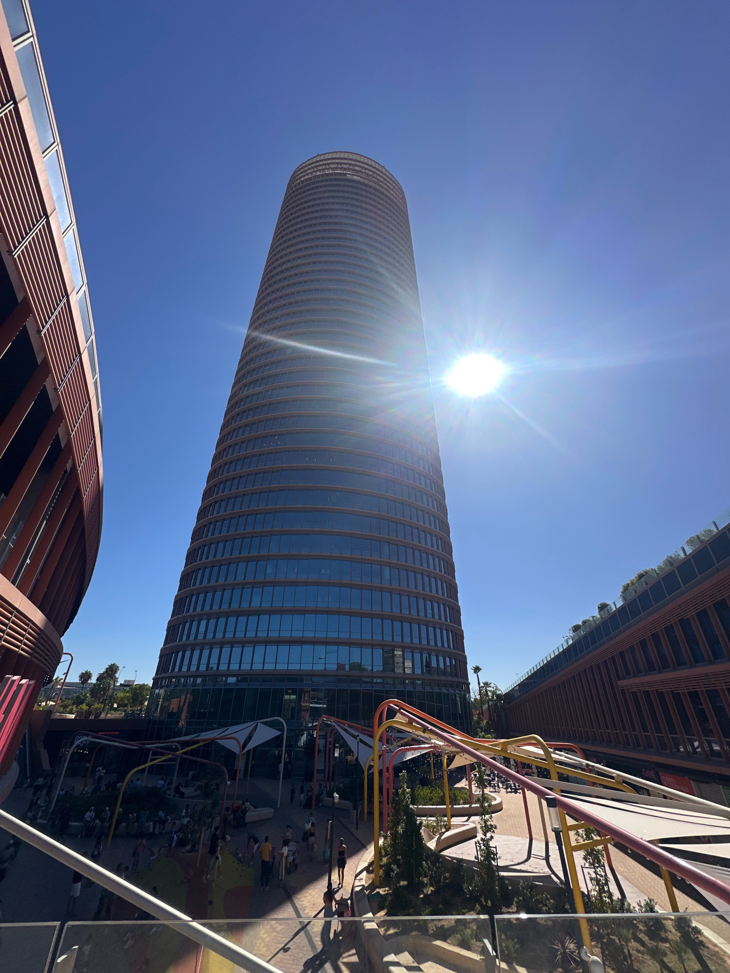 The image depicts the Metropol Parasol, also known as "Las Setas" (The Mushrooms), in Seville, Spain. The iconic wooden structure with its mushroom-like caps dominates the skyline, with the Torre Pelli, a prominent skyscraper, visible in the background. The sky is a clear, bright blue with the sun shining intensely, causing lens flare.
In the foreground, people are seen walking and congregating in a public space beneath the Metropol Parasol. The area appears to be a plaza or a pedestrian walkway, with several colorful, modern structures and seating areas. There are also some trees and landscaping elements. The architecture of the surrounding buildings is modern and sleek, with large glass windows and warm-toned facades. The overall scene suggests a vibrant and active urban environment on a sunny day.