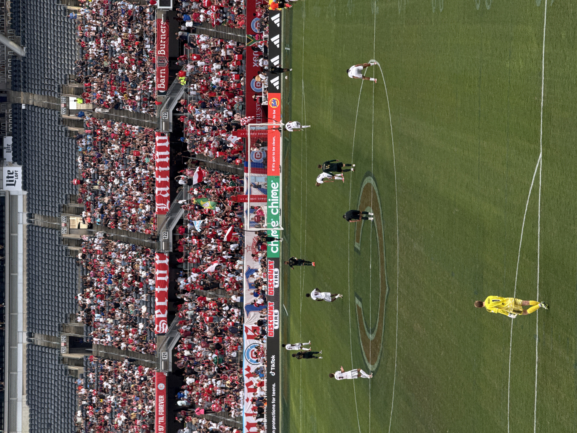 This is a high-angle, full shot of a professional soccer game in progress at Soldier Field in Chicago, Illinois, United States of America. The field is green with white lines marking the boundaries and penalty areas. Several players in white and dark uniforms are scattered across the field, some in motion and others standing. One player in a bright yellow goalkeeper's uniform is positioned on the left side of the frame. In the background, a large crowd of spectators, predominantly dressed in red, is seated in the stadium's tiered seating. Banners with various sponsorships and text are visible along the stadium's upper levels. The lighting suggests it is daytime.