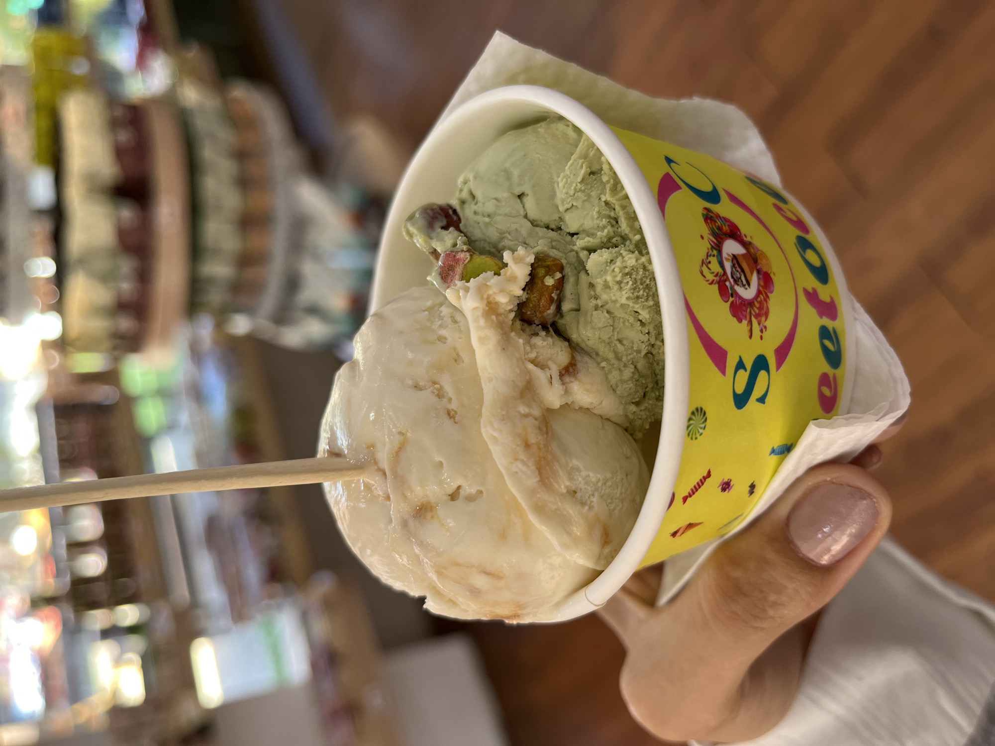 A close-up shot shows a person holding a cup of ice cream. The ice cream is in a yellow and white paper cup with a colorful logo. There are two flavors of ice cream visible: one is a light green color and the other is a light tan or off-white color with brown swirls. The ice cream is garnished with chopped pistachios. A wooden stick is stuck into the ice cream. The person holding the ice cream cup has pale skin and manicured nails. The background is blurred, but appears to be a store with shelves of items. The location is Unknown, Indonesia.