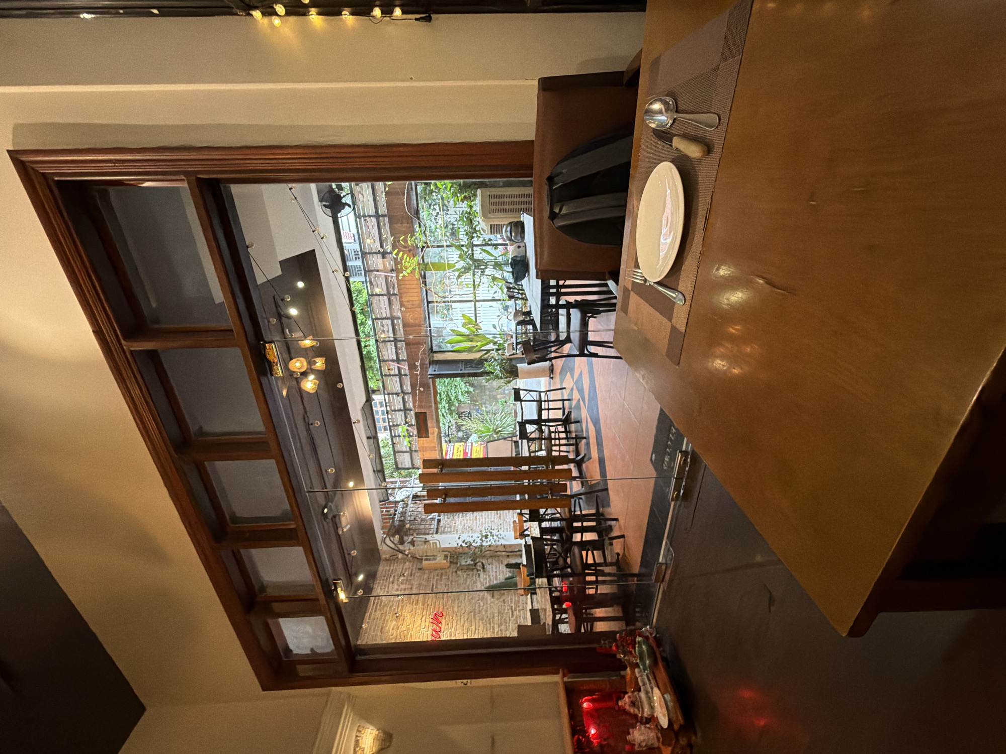 This is a medium shot, eye-level photograph taken indoors, looking through a glass doorway into a restaurant's outdoor seating area. The foreground features a wooden table with a place setting including a white plate, fork, knife, and spoon on a placemat. A brown leather chair is partially visible behind the table, with a black backpack resting on its back.
The doorway leads into a covered outdoor courtyard or patio, visible through the glass. This area contains multiple tables and chairs, some with diners, though their features are not discernible. The walls in the courtyard are exposed brick, and there are plants, lighting, and a railing visible. The overall impression is of a cozy, possibly mid-range restaurant. The context provided indicates the location is the city of Thủ Đức, Vietnam.
