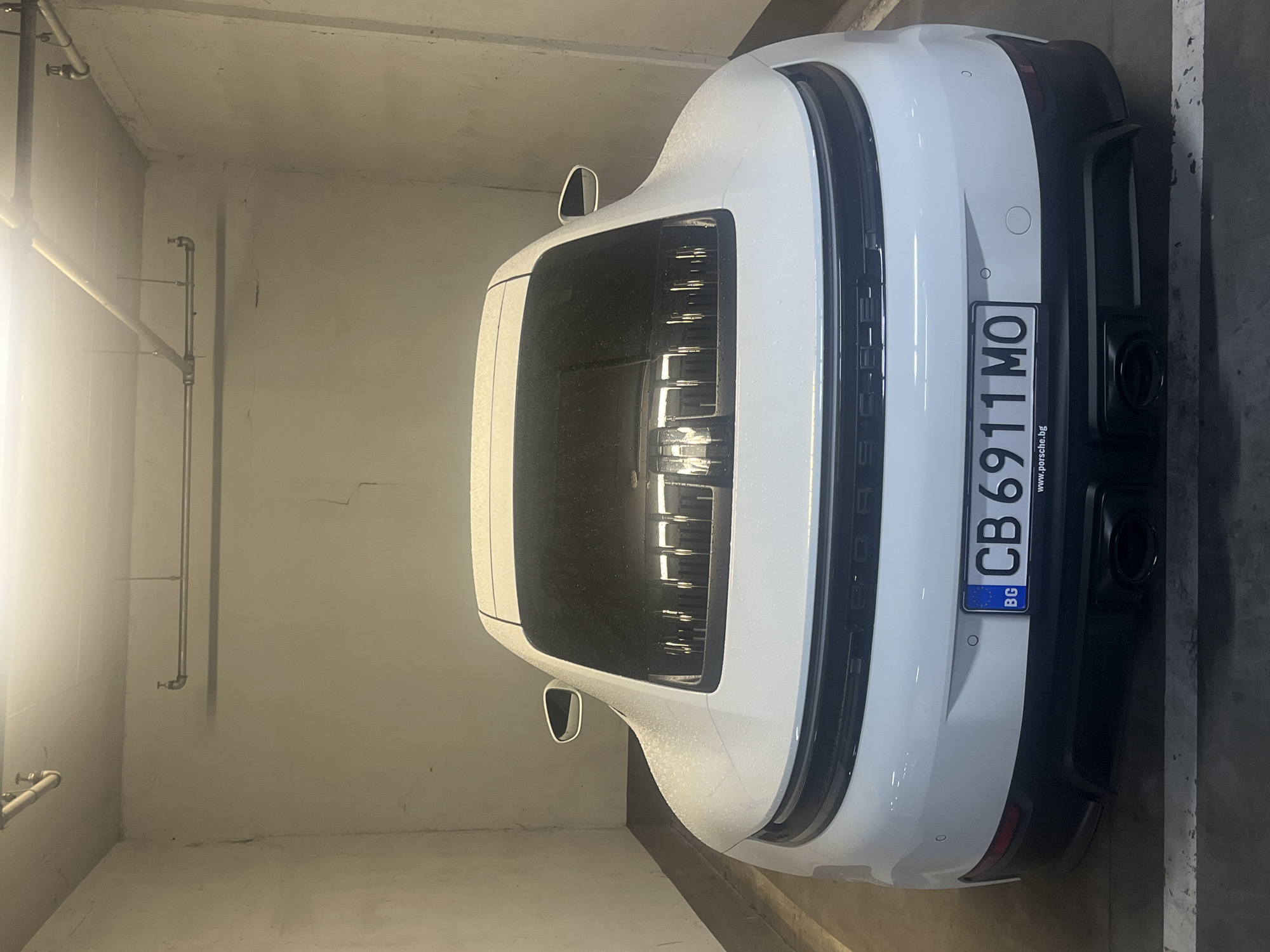 A white Porsche 911, bearing Bulgarian license plate CB 6911 MO, is parked in an indoor parking garage. The location is likely within Sofia, Bulgaria, given the license plate's "BG" country code. The car is positioned close to a light-colored wall with visible piping. The rear of the vehicle is facing the viewer.