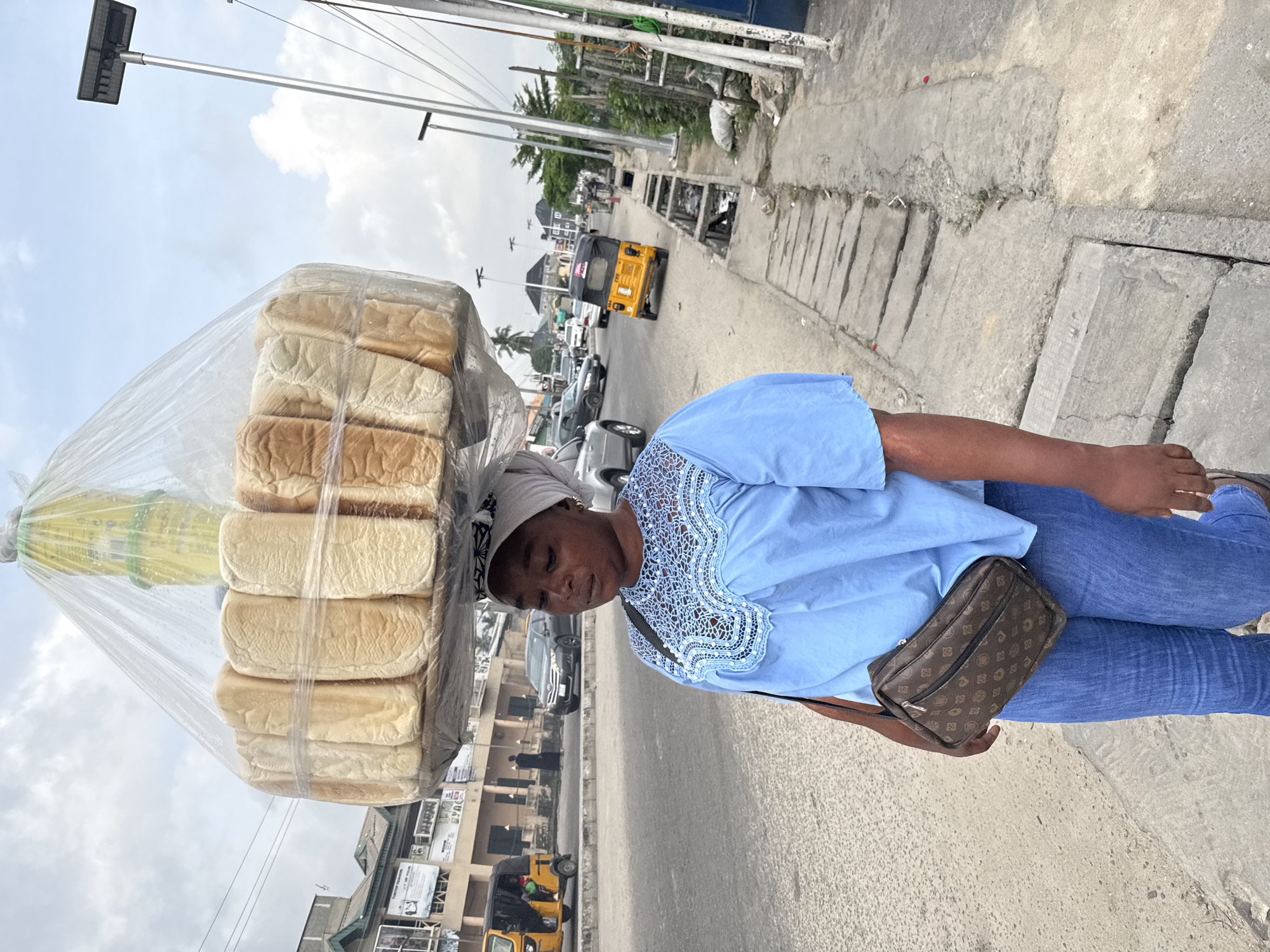 A woman is seen carrying a large stack of loaves of bread on her head, wrapped in a clear plastic bag. She is walking on a sidewalk in Ado, Nigeria. The woman is wearing a light blue blouse with lace detailing around the collar, blue jeans, and a white head covering. She has a brown purse slung across her shoulder. In the background, there is a street with cars, a yellow auto rickshaw ("Keke"), and buildings. A streetlight pole is visible above. The sidewalk is concrete. The sky is overcast.