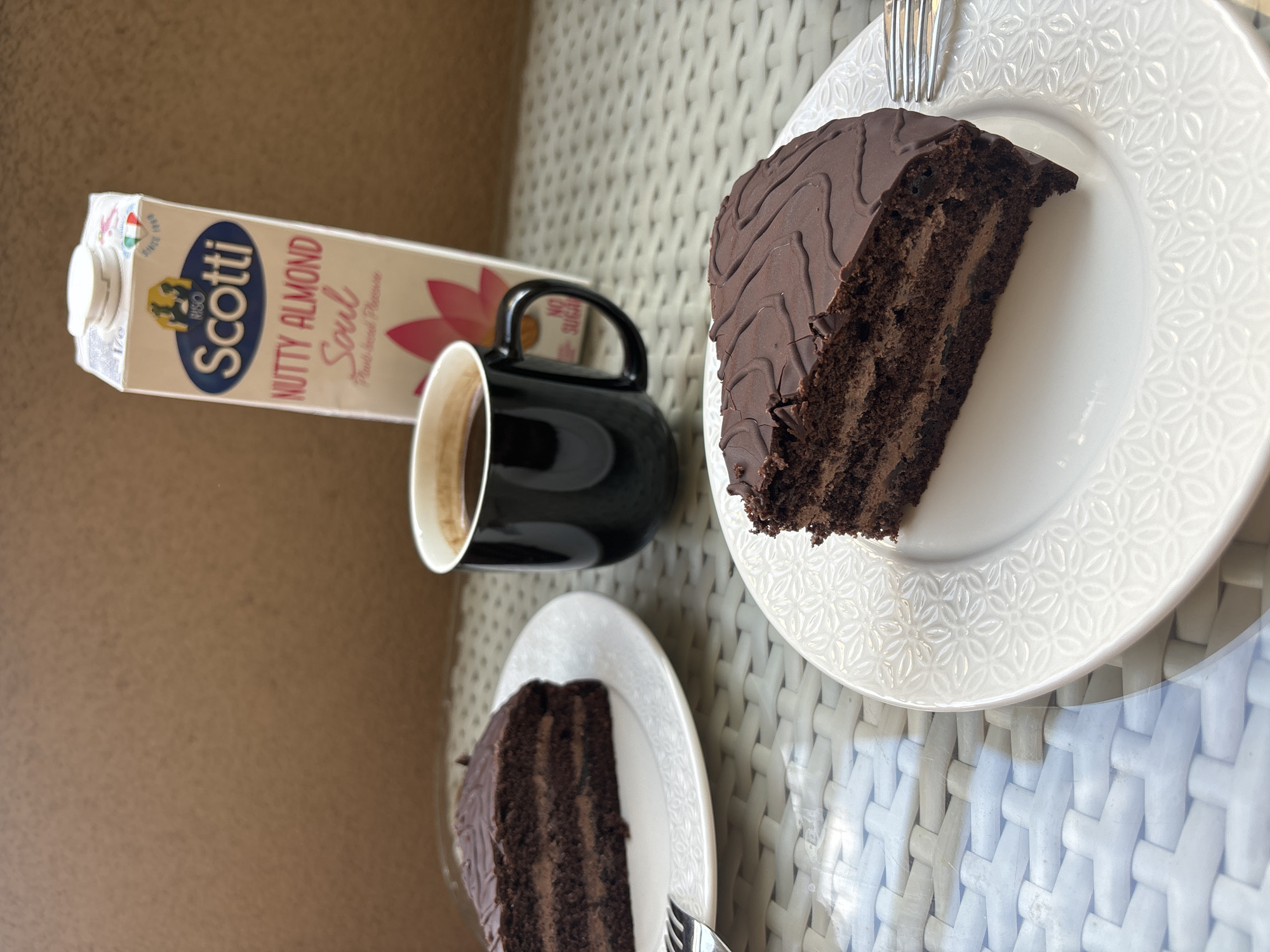 A close-up, eye-level shot shows two slices of chocolate cake and a black mug of coffee on a textured, light-colored table outdoors. To the right, a carton of Scotti Nutty Almond Soul Plant-based drink is visible against a brown, textured background. The slice of cake in the foreground is on a white plate with a floral embossed pattern and has a decorative chocolate drizzle on top. A silver fork rests to the right of this plate. A second slice of cake is on a similar white plate to the left, slightly out of focus, with another fork resting on it. The coffee mug is black with a white interior and contains dark coffee. The lighting suggests a bright, possibly sunny, outdoor environment.