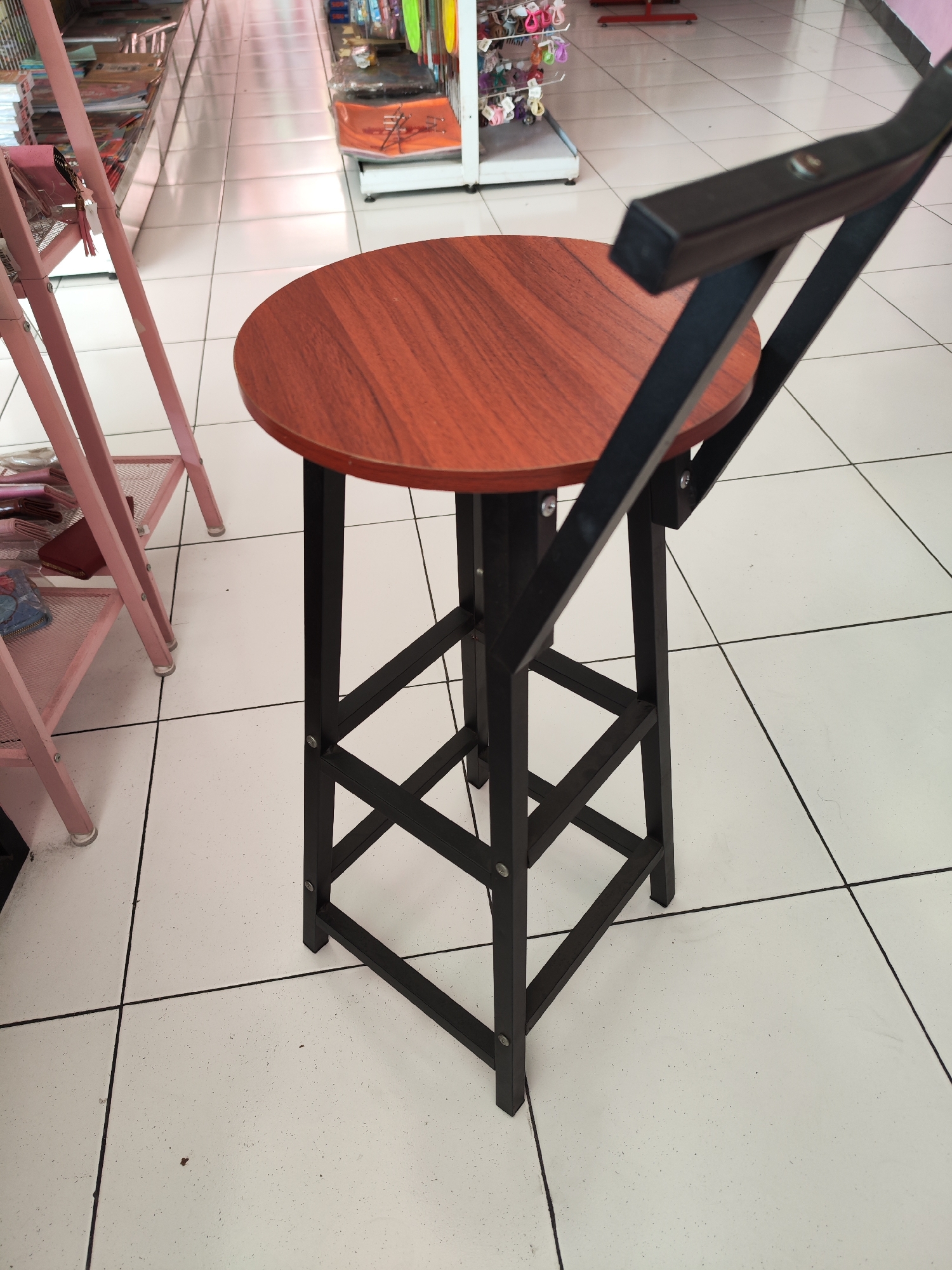 A dark brown, round bar stool with a black metal frame stands on a white tiled floor. The stool has a wooden seat and a black metal backrest. The floor is made of large white tiles with black grout lines. In the background, there are shelves stocked with various items, suggesting a retail environment. To the left of the stool, there is a pink metal shelf unit. The lighting in the image is bright and even.