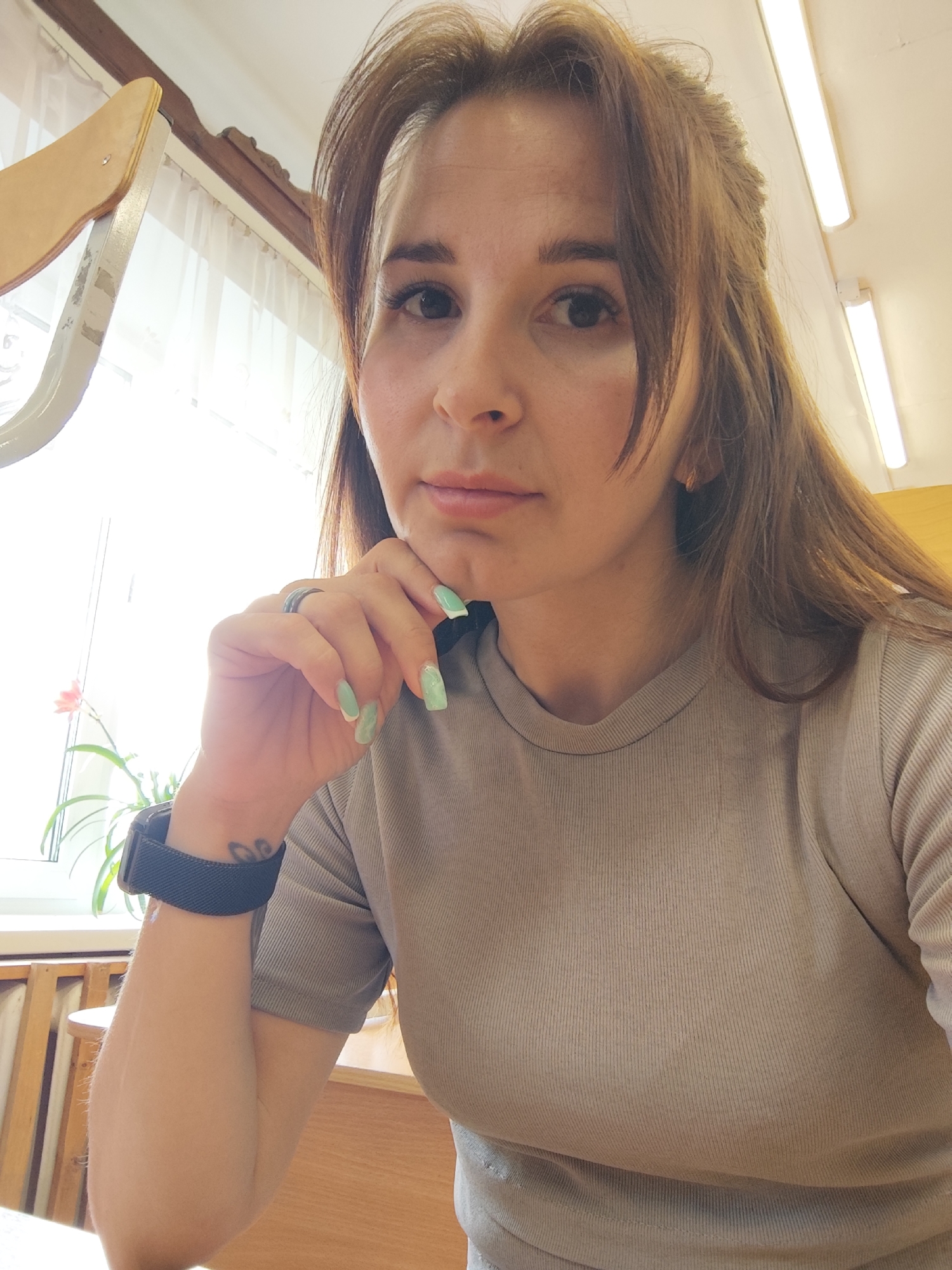 A woman with brown hair and light skin is sitting indoors. She wears a gray shirt, a black watch on her left wrist, and a ring on her left hand. Her left hand is raised to her face, with her fingers near her chin. In the background, there is a window with sheer white curtains, a green plant on the windowsill, and fluorescent lights on the ceiling. Wooden furniture is visible. There is no direct evidence to indicate the exact location within the city of Фоки, Russia.