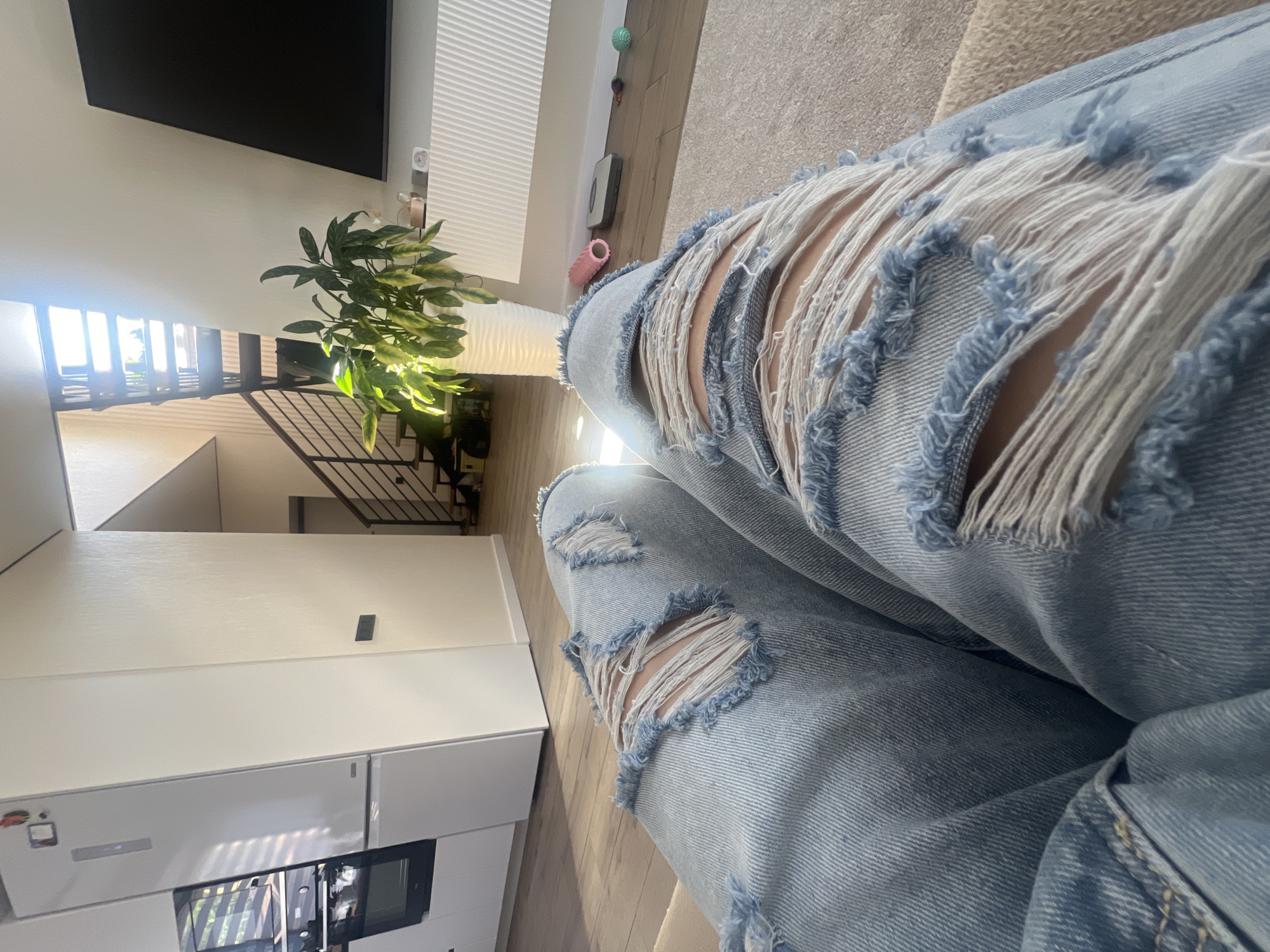 This is a first-person perspective, slightly tilted shot of a person wearing distressed denim jeans. The jeans are light blue with prominent rips, revealing skin underneath. The background suggests an indoor setting, possibly a modern living room or entryway. To the left, a sleek white cabinet unit is visible, with what appears to be a refrigerator and an oven or microwave integrated. Above this, a staircase with dark metal railings leads upwards, and a large plant with variegated leaves is situated near the stairs. Further up and to the left, a black rectangle, likely a television, is mounted on a light-colored wall. The wall itself has a textured finish and is partially illuminated by sunlight filtering through blinds, creating striped patterns of light and shadow. In the upper right portion of the frame, a carpeted floor is visible, along with a hint of a light-colored sofa. The overall lighting suggests daytime. There is no visible text in the image. The scene is static, with no specific activity clearly depicted beyond the subject's presence.