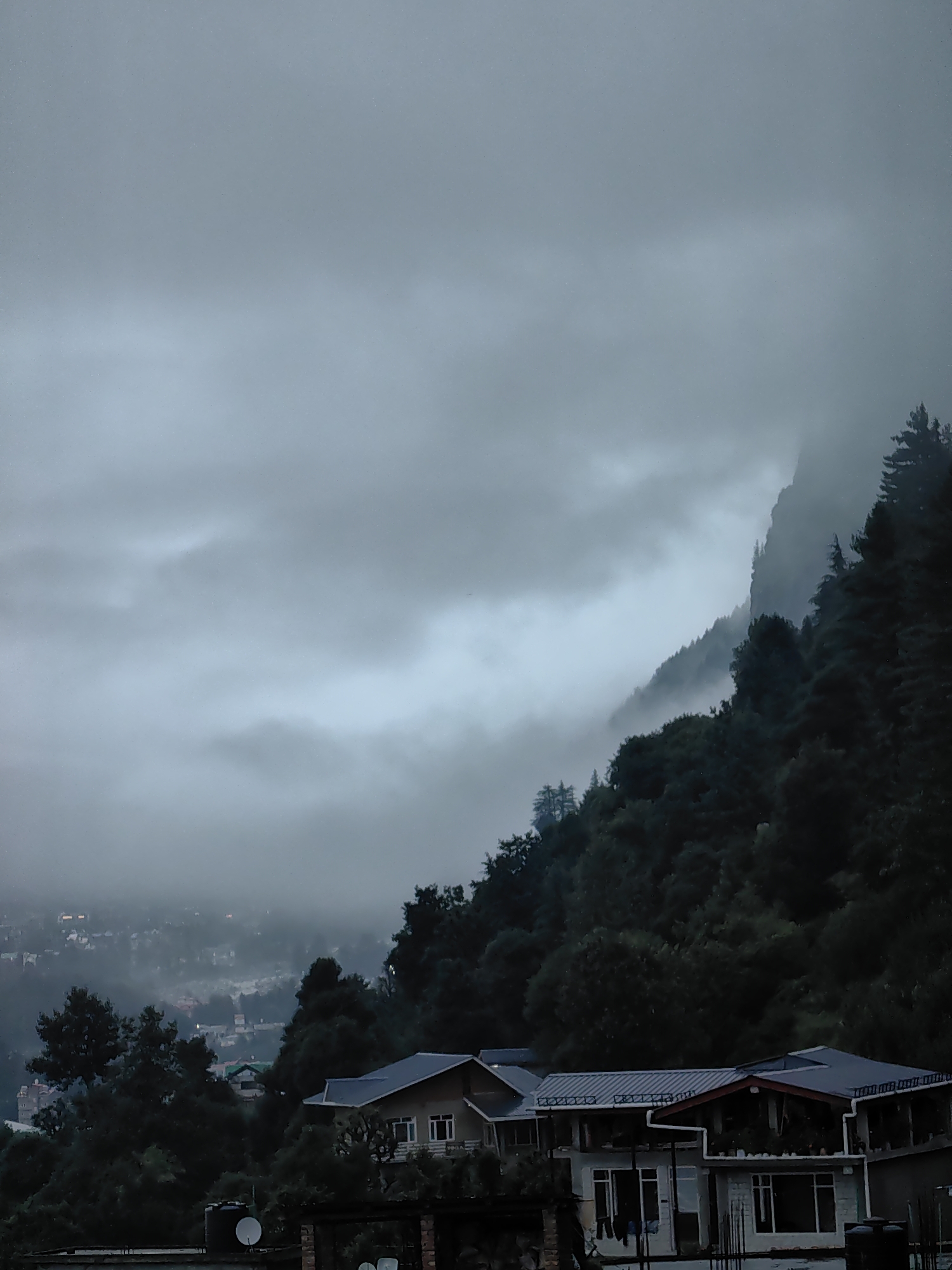 The image captures a view of Vashisht, India, on a cloudy day. The foreground features houses with dark roofs, partially obscured by trees. In the mid-ground, more houses with lights are visible through the mist, forming a village scene. The background is dominated by a steep, mist-shrouded mountain covered in dense green trees, with clouds obscuring the upper reaches of the mountain. The overall atmosphere is muted and overcast.