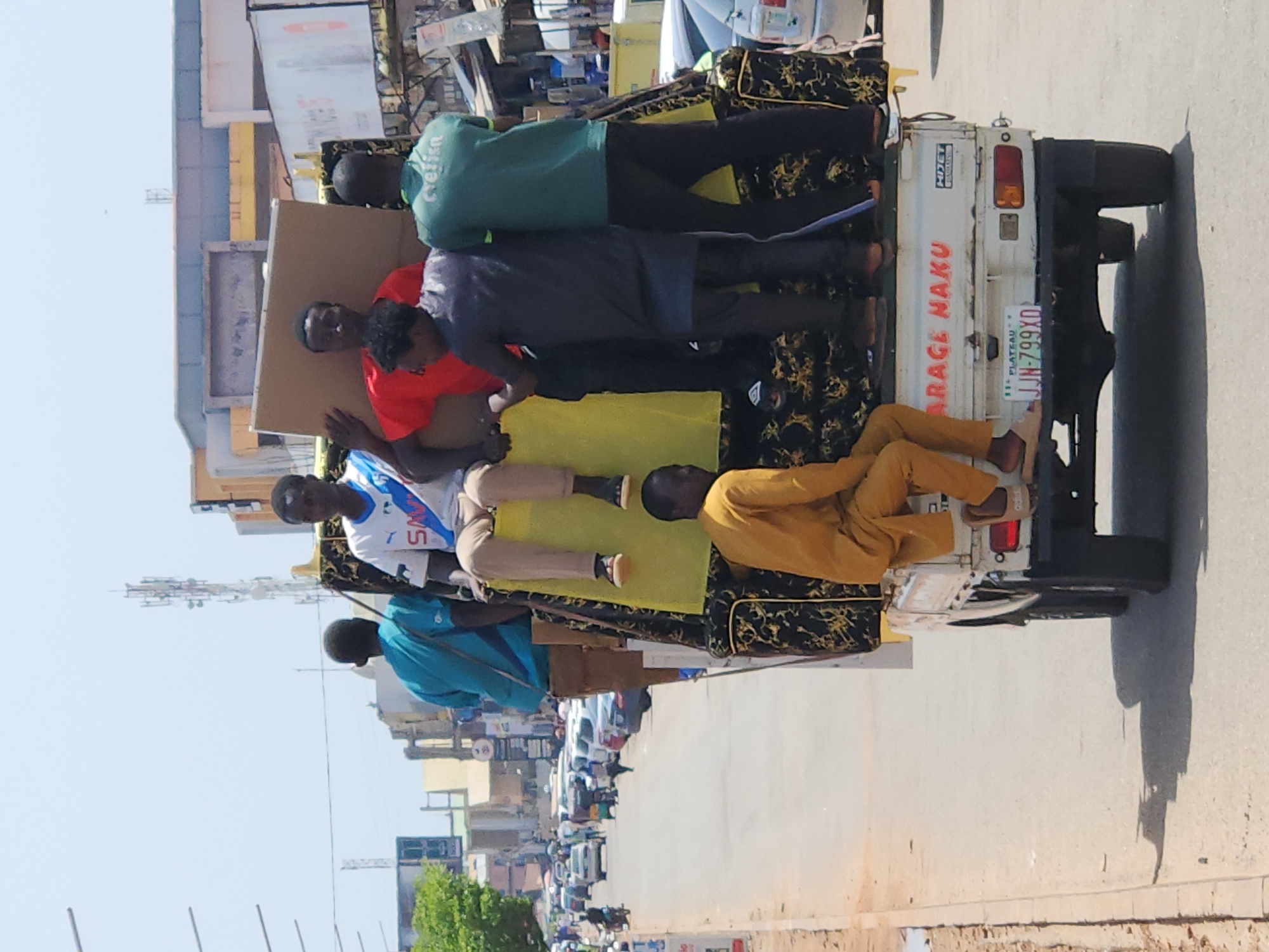 A group of six young men are standing and sitting on top of a truck in a city street. The truck is loaded with furniture, including what appears to be a sofa covered in a patterned fabric and a large piece of cardboard. The men are dressed in casual clothing, with some wearing t-shirts and others in traditional attire. One man in the front is wearing a yellow outfit, while another in red holds the cardboard. The truck is driving on a paved road with cars and motorcycles in the background, suggesting a busy urban environment. The sky is clear and bright, indicating a sunny day. The license plate on the truck reads "JJN-799XD" and indicates the location as "PLATEAU". The text "ARAGE NAKU" is visible in red on the back of the truck. This scene is consistent with a typical transportation of goods and people in the city of Jos, Nigeria.