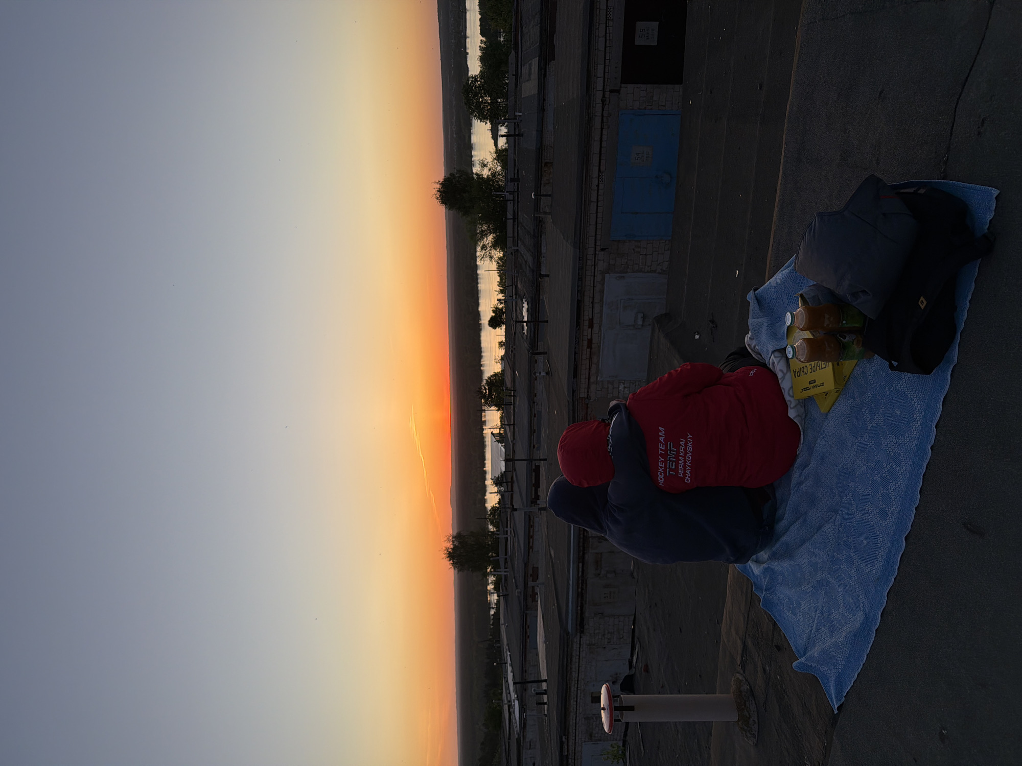 Two individuals sit on a light blue blanket laid on a rooftop at sunset. One wears a red jacket with "HOCKEY TEAM TENTIF PERM KRAI CHAYKOVSKIY" printed on the back. A yellow box with four bottles sits on the blanket. Beside the blanket are two folded jackets. The rooftop overlooks a river and cityscape at sunset in Chaikovsky, Russia. The river runs along the horizon. In the middle ground are the tops of trees and buildings. In the background, the sky transitions from orange to gray.