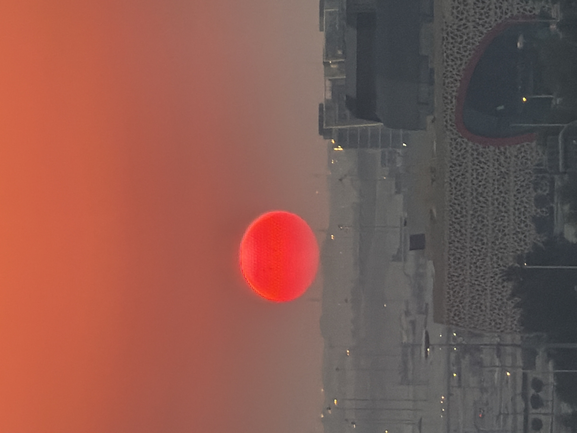 An extremely close-up, vertical, eye-level shot captures a large, vibrant red sun low in a hazy orange sky. The sun, appearing almost perfectly spherical, is the dominant subject, its bright red hue contrasting with the muted tones of the background. Below the sun, a cityscape is visible, rendered in soft focus and a greyish hue due to atmospheric conditions. Various buildings, some appearing modern with straight lines and others more textured, are discernible. Streetlights dot the landscape, their yellow glow appearing as small points of light. The foreground features a distinctive building with a patterned facade and an arched entrance, its details softened by the distance and haze. The overall impression is of a sunset or sunrise over an urban area, with the dense atmosphere creating a diffused and serene ambiance.