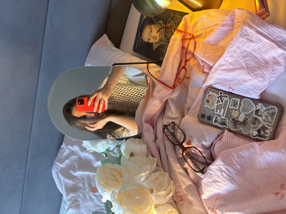 The image depicts a bedroom scene, likely in an apartment or hotel room within a Russian-speaking country, but the specific location within Krasnodar, Russia, cannot be determined from visual information.
In the foreground, a bed is visible with a grey upholstered headboard. The bedding consists of a white fitted sheet and a light pink duvet cover featuring a faint white stripe pattern and small red bow motifs. On the bed, from left to right:
1. A bouquet of artificial white or cream-colored roses rests on the duvet.
2. A pair of tortoiseshell-patterned reading glasses is placed on the duvet.
3. A silver iPhone, identifiable as an iPhone 11 due to its camera array, in a clear case, lies face up on the duvet. The case is extensively decorated with numerous vintage-style stickers, including images of cherubs, a Roman bust, dogs, and text snippets in English and Russian (e.g., "my darling all i want is to love you," "МЫ"). An Apple logo sticker is also present.
4. A pair of red-framed reading glasses is positioned on the duvet towards the right.
5. Partially visible on a bedside table to the right is a dark hardback book titled "ЭЛИСОН УЭЙР" (Alison Weir), featuring a historical portrait of a young woman (likely a depiction of Queen Elizabeth I) on its cover. A metallic, gold-toned object, possibly the base of a lamp, is also present next to the book.
Dominating the center of the image is an oval-shaped tabletop mirror with a black stand, positioned on the bed. Reflected in the mirror is an adult female. She has long, dark hair and is wearing a light-colored, open-knit or mesh top. Her left hand is held near her face, and her right hand is extended, holding a red-cased smartphone, indicating she is taking a mirror selfie. A ring is visible on her left ring finger, and a delicate bracelet is on her left wrist. The overall scene appears to be an intimate, personal bedroom setting, illuminated by soft, possibly natural light.
