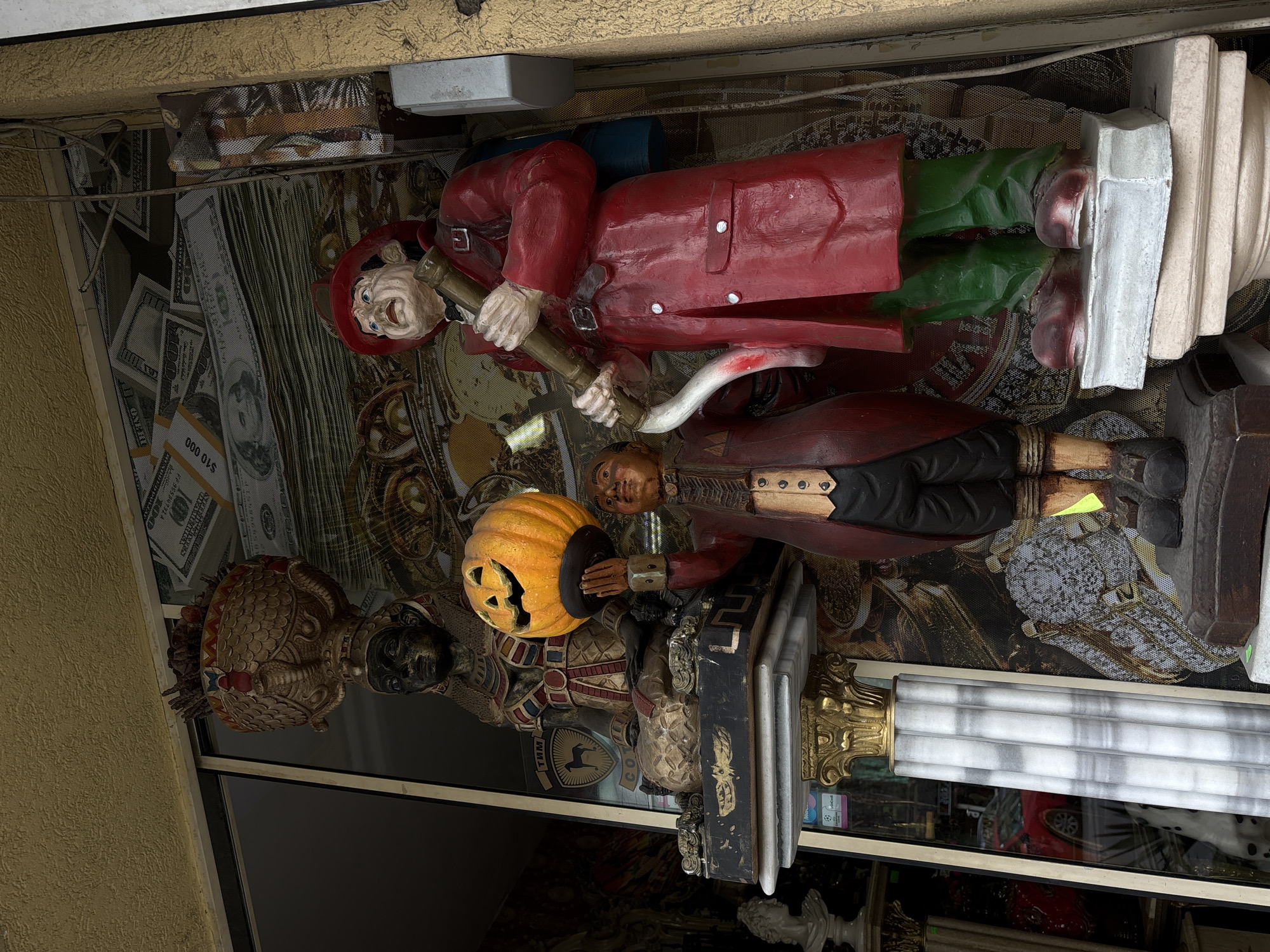An eclectic and colorful display of decorative figures is showcased in what appears to be a shop window, possibly in Varna, Bulgaria. The scene is dominated by three large statues. On the right, a cartoonish figure, perhaps a pirate or an adventurer, stands tall, dressed in a bright red trench coat, green trousers, and oversized red shoes. He wears a red hat and sports an exaggerated smiling face with blue eyes, holding a brass-colored telescope or pipe. His feet rest on a white, architectural base.
Next to this figure, a darker-skinned statue, clad in a historical-style coat, holds a bright orange, carved jack-o'-lantern, suggesting a Halloween or autumn theme. To the far left, another figure, resembling an ancient warrior or deity, is depicted with dark skin, intricate golden-bronze armor, and a textured helmet or headdress. The letters "TMM" are visible on its chest.
The background behind these statues features a busy, patterned screen or wallpaper. It is adorned with numerous stacks of US hundred-dollar bills, some clearly marked "$10 000," interspersed with images of clocks and other ornate designs. A metallic mesh screen is also visible in the background. To the far left, a textured yellow wall frames the display. On the right side, a prominent white fluted column with a golden capital adds a classical touch. The lower part of the display features a dark, reflective surface and hints of other decorative items, including a white classical bust. The overall display is brightly lit, creating a vibrant and varied visual experience.