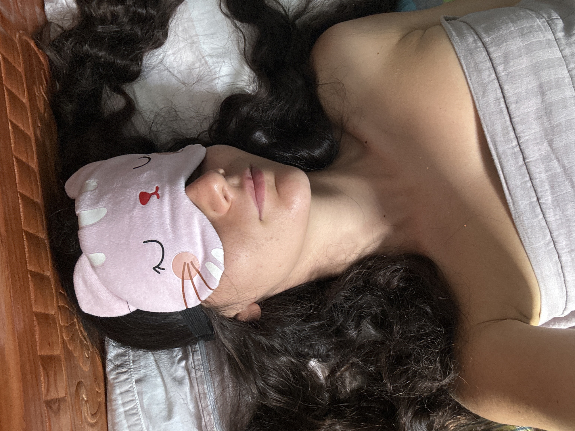 A woman is lying on her side on a bed with her head facing left. She is wearing a grey tank top and a pink cat-shaped eye mask. Her dark, curly hair is spread around her head on a white pillow. The bed is next to a wooden bed frame. The location is likely Mũi Né, Vietnam.