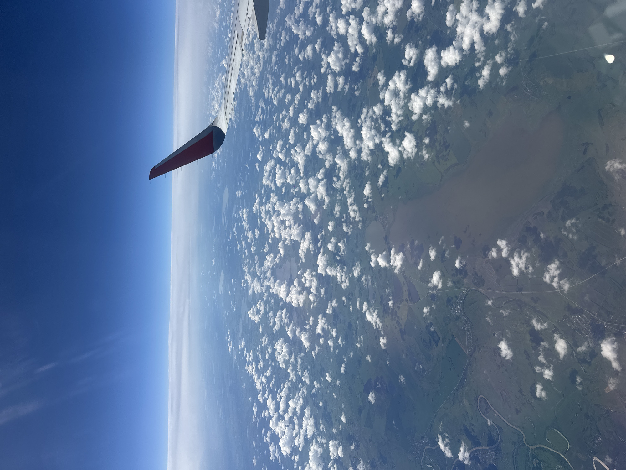 The image is taken from an airplane window, looking down at a vast landscape below. The perspective is tilted, with the airplane wing prominently visible in the foreground on the left side of the frame. The wing is white with a red and black tip, suggesting it's part of a commercial aircraft.
Below the wing, the scene is divided into distinct layers. The upper left is dominated by a deep, clear blue sky, transitioning into a hazy, lighter blue near the horizon. A thin band of white clouds stretches across the horizon.
The majority of the image is a view of the earth from high altitude, dotted with numerous fluffy white cumulus clouds casting shadows on the land below. The landscape beneath the clouds is a patchwork of green fields, possibly farmland, interspersed with numerous small bodies of water, likely lakes or ponds. A prominent, winding river with darker water flows through the scene, with tributaries branching off. There are signs of human habitation, with small clusters of buildings visible, suggesting villages or towns. Roads and pathways crisscross the terrain. The overall impression is one of a rural, possibly somewhat remote, location.
The lighting suggests it is daytime, with the sun high enough to cast clear shadows from the clouds. The weather appears fair, with scattered clouds and clear patches. There is no visible text in the image. Given the location context provided as Ragozina, Russia, this depicts a view over that general region. The activity is passive observation from an aircraft in flight.