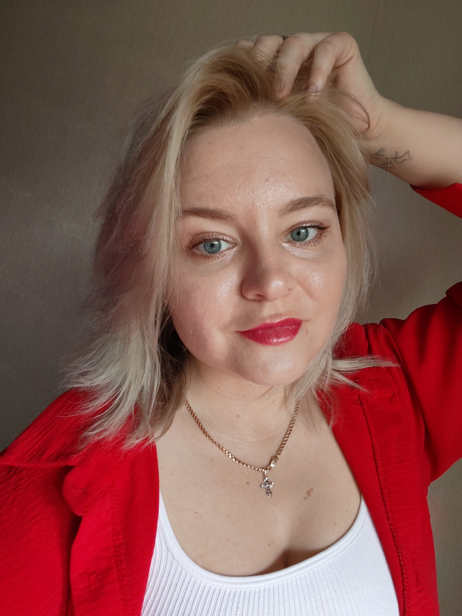 A light-skinned woman with blond hair and blue eyes is seen from the chest up in a selfie-style shot. She has red lipstick on. Her right hand is touching her hair. She is wearing a red shirt over a white tank top and a gold necklace with a cross pendant. A tattoo is visible on her right forearm. The background appears to be a plain, light-colored wall. The location in Izhevsk, Russia is not discernible from the image.