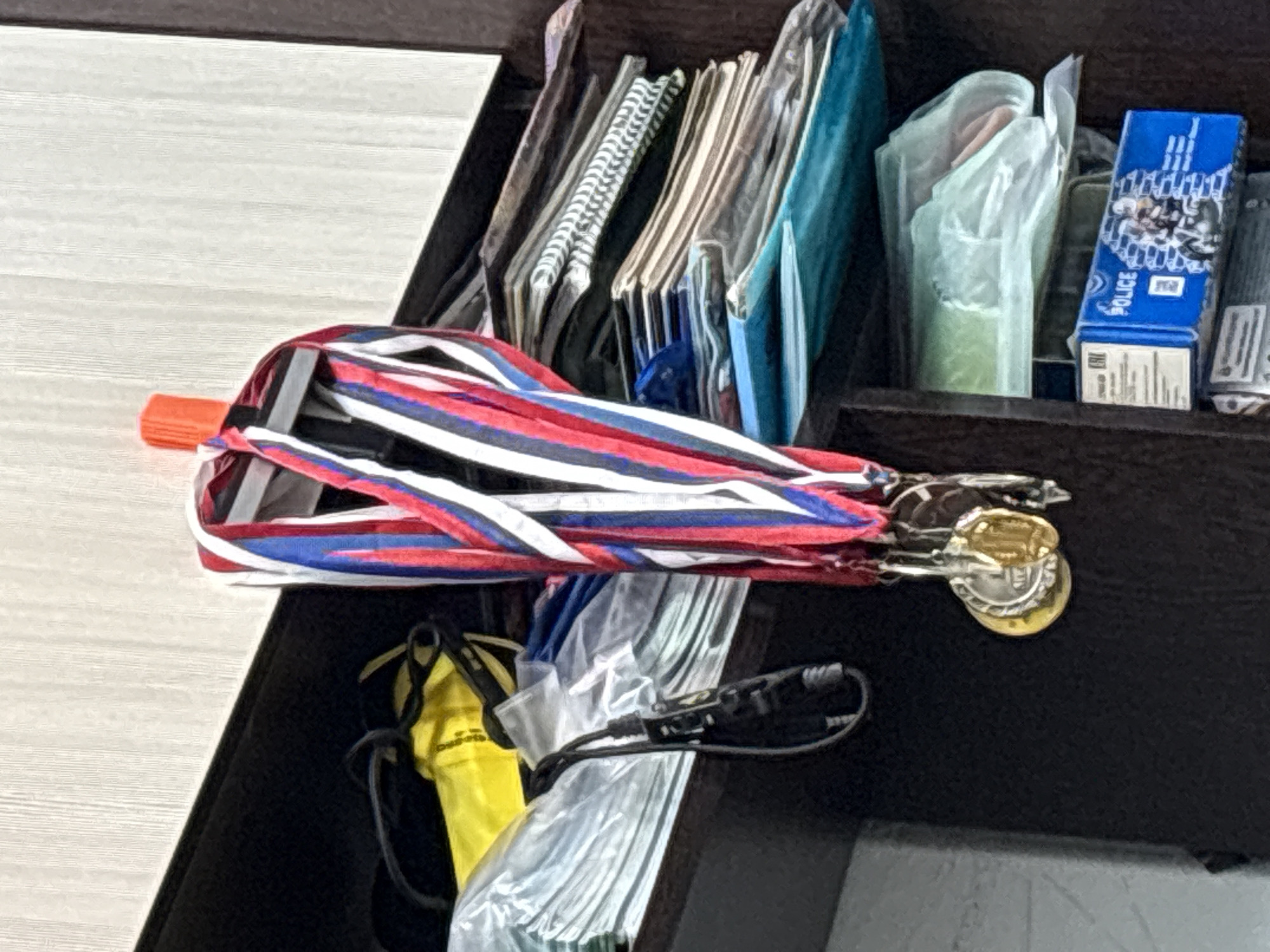 A close-up shot shows medals hanging from a small orange hook on a shelf in an interior, likely a closet or storage area. The medals have red, white, and blue ribbons. Below the medals, there are various stacked items, including what appear to be folders, notebooks, and plastic-wrapped objects. A blue box with the word "POLICE" and an image of a motorcycle and figures is visible on the bottom shelf. The background wall has a textured, light-colored surface, and the shelf is dark.