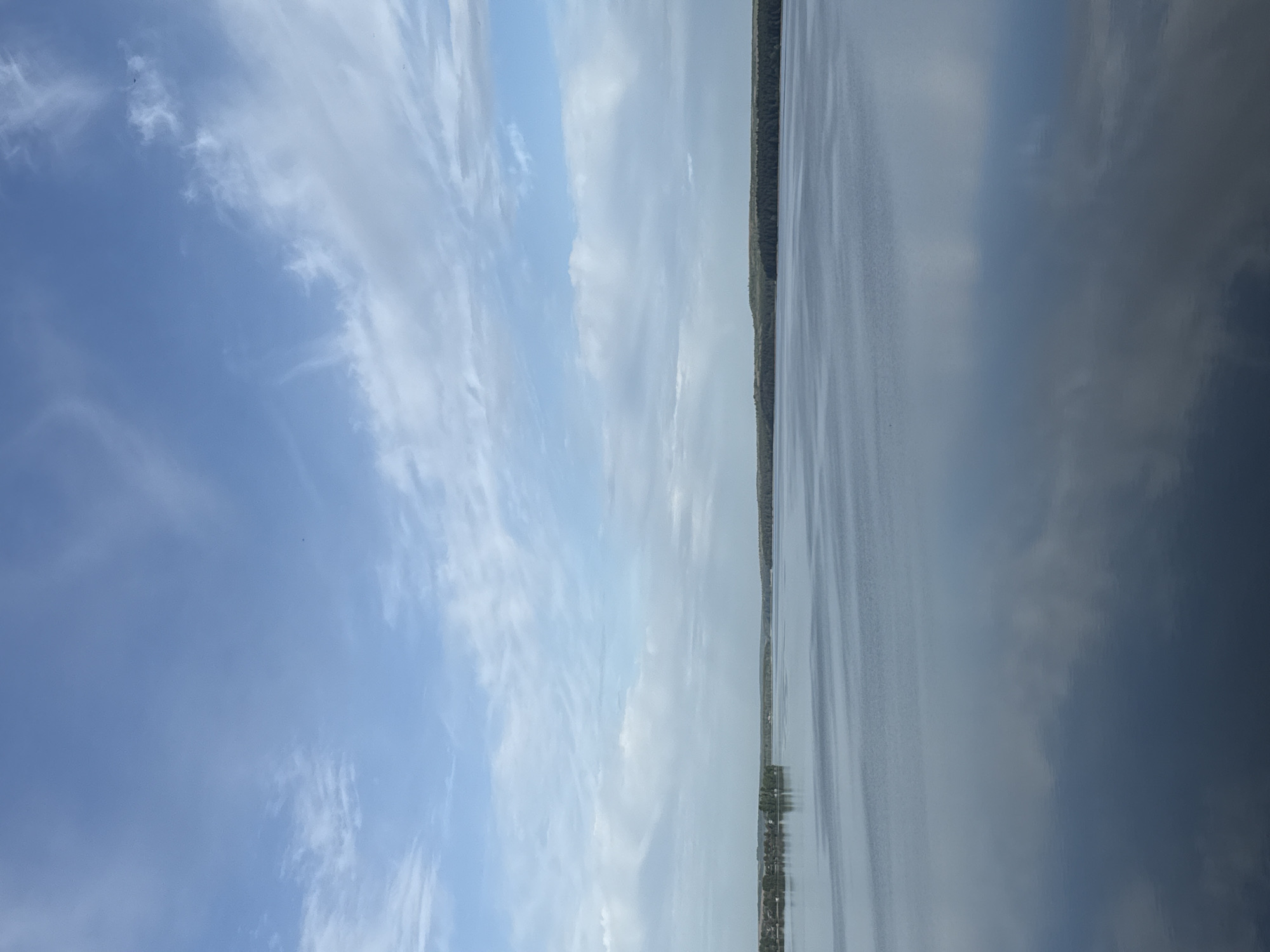 The image depicts a serene lake scene near Votkinsk, Russia. The left side of the frame showcases a bright, blue sky filled with wispy clouds. A thin strip of land with trees runs horizontally through the middle of the image. The right side shows the lake, mirroring the sky and clouds above, creating a symmetrical effect. The water surface is mostly still, showing gentle ripples. There are no visible people or objects.