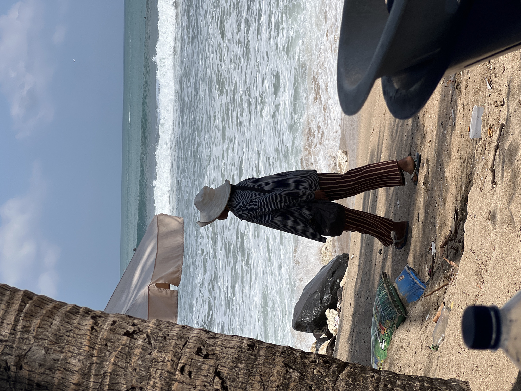 A person wearing a wide-brimmed hat, a striped shirt, and striped pants walks on a sandy beach in Kuta, Indonesia. The person carries a dark bag over their shoulder. Waves from the ocean are visible behind them, with a few surfers in the water. A palm tree trunk is in the foreground on the left, and a large dark rock is on the sand. Several pieces of trash, including plastic bottles and bags, are scattered on the beach. In the lower right corner, a dark, curved object, possibly part of a bin or container, is visible. The sky is blue with some clouds, and a faint airplane can be seen in the distance.