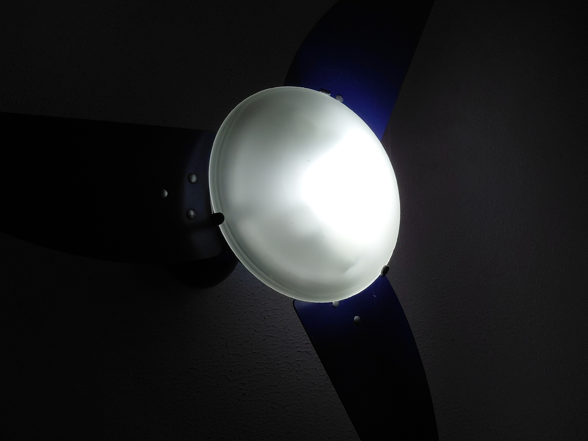 The image depicts a ceiling fan with three blades. The blades appear to be dark in color, possibly black or dark blue. A light fixture is centered on the fan, emitting a bright, diffused white light. The light illuminates the fan blades and the surrounding area, which seems to be a ceiling with a textured surface. The overall scene is dimly lit, with the light fixture being the primary source of illumination. There is no indication of the location being Santa Ana, Venezuela, or any people or actions taking place.