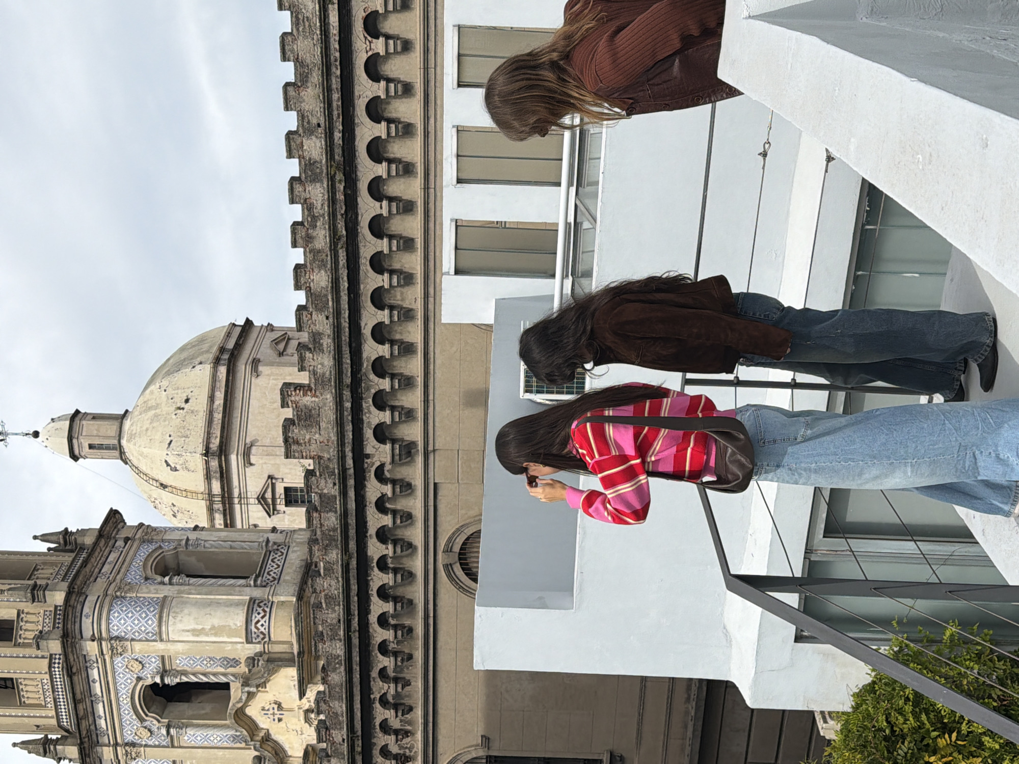 Three women are standing on a rooftop, observing and photographing a historic building. The building features a dome, a tower adorned with decorative blue tiles, and ornate stonework. The women are positioned along a white railing with a black metal fence. One of the women is actively taking a picture with a phone or small camera. The scene is set in Buenos Aires, Argentina, indicated by the architectural style. The weather appears overcast, with a light gray sky.