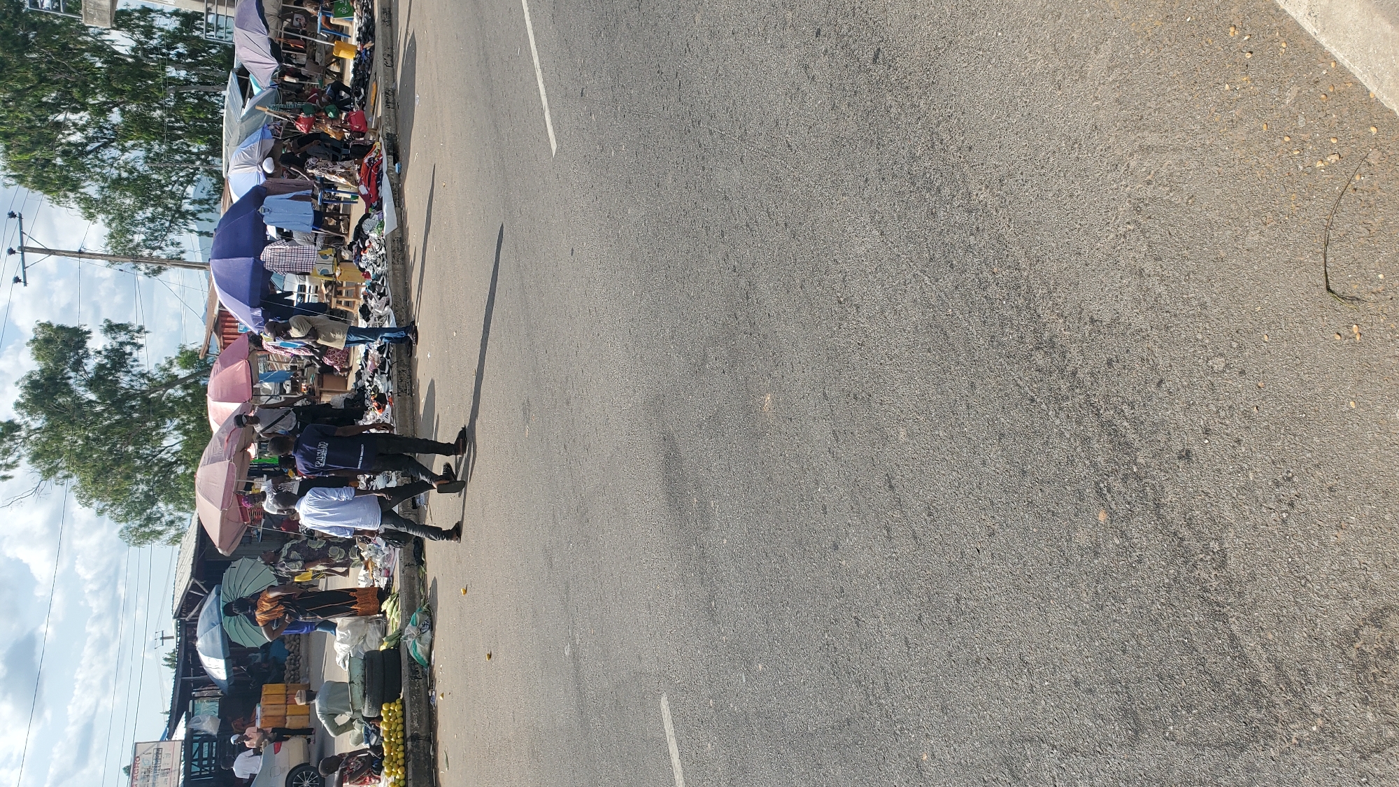 The image shows a street market scene in Jalingo, Nigeria, on a sunny day. Several people are present, some browsing merchandise displayed under umbrellas and makeshift stalls. The merchandise appears to be primarily clothing and shoes, laid out on the ground and on tables. Two men in the foreground are walking away from the camera. A car is visible on the left side of the image, and trees and power lines are in the background. The market is set up along the side of a paved road, with a concrete curb separating the market area from the road. Yellow containers are stacked on the left side of the market. The overall impression is of a bustling, informal marketplace.
