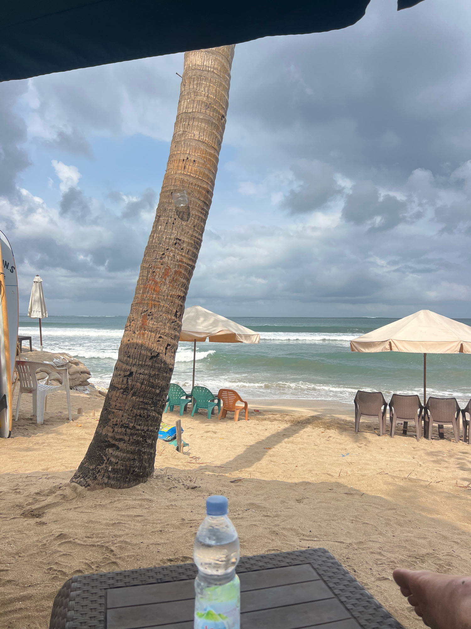 A scene on a beach in Kuta, Indonesia, shows a sandy shore with a large palm tree dominating the foreground. The tree trunk is thick and textured, leaning diagonally across the frame from bottom left to upper right. In the background, the ocean is visible with moderate waves and a cloudy sky. Several beach chairs and umbrellas are set up on the sand, suggesting a leisure area. On the left side, a surfboard leans against something unseen. A plastic bottle of water sits on a dark, textured table in the lower right foreground. There are no people clearly visible in the scene, only a foot in the extreme bottom right corner. The overall atmosphere is that of a cloudy day at the beach.