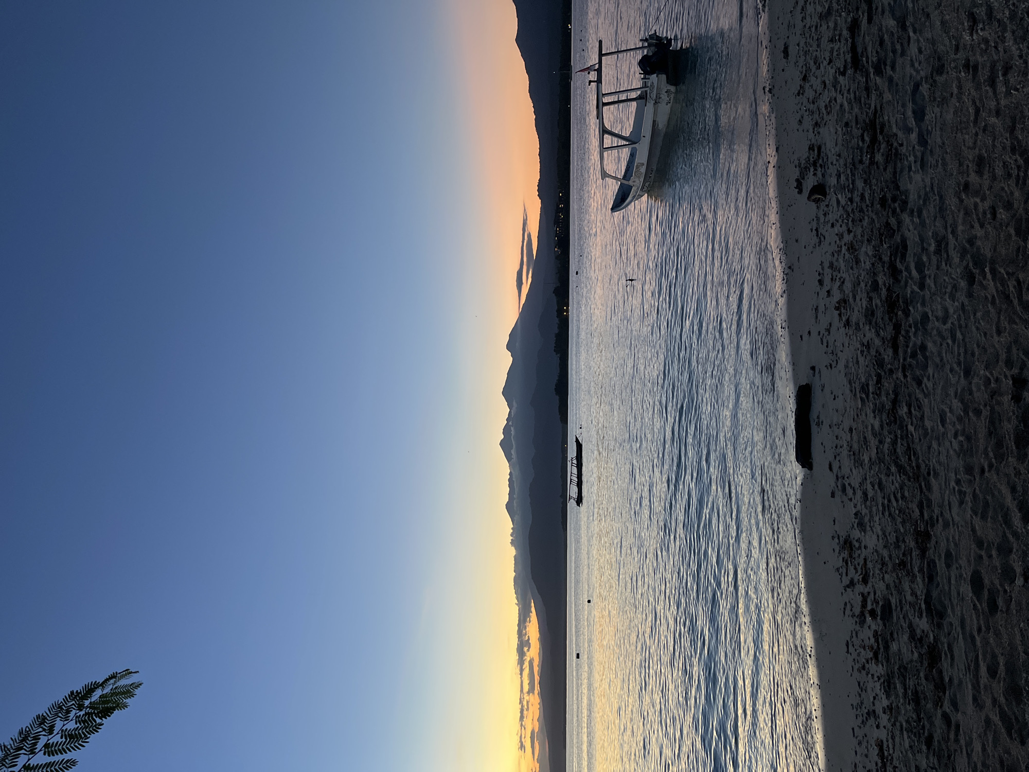 A horizontal image depicts a beach at dusk. The sky transitions from dark blue at the top left to orange on the horizon. A mountain range is silhouetted against the sunset. The ocean is textured with ripples and reflects the sky's colors. Two boats are anchored offshore. The closest boat is white with an outboard motor. The beach is dark sand. A leafy branch is visible in the lower left corner. The location is Unknown, Indonesia.