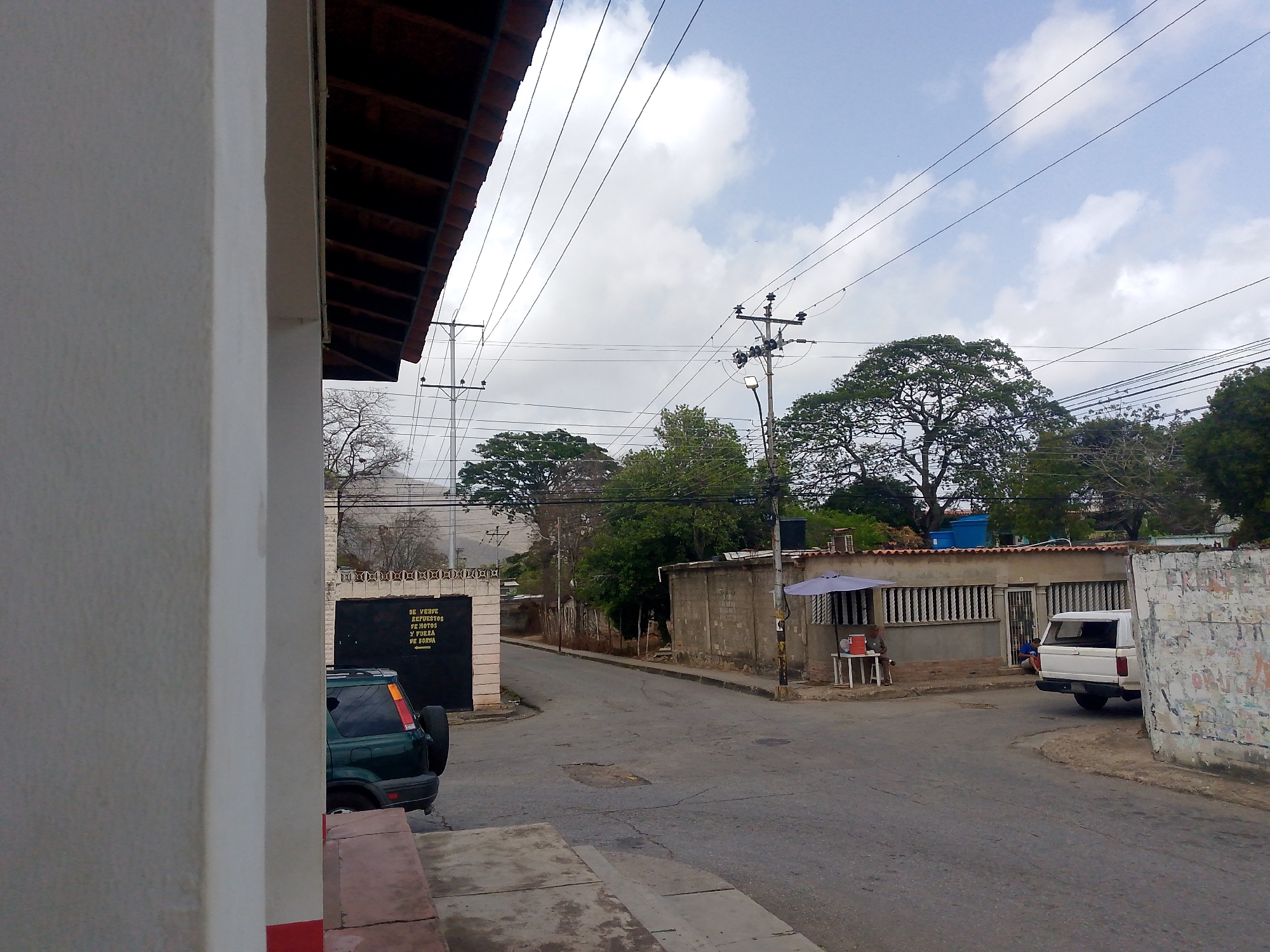 This media file captures a daytime street scene in Santa Ana, Venezuela, under a partly cloudy sky. The image is framed on the left by a white building wall and the underside of a terracotta-tiled roof.
The scene depicts a quiet, paved road that gently curves to the right. On the left side of the street, a dark green SUV with a spare tire mounted on the back is parked partially visible, behind a large black gate. On this gate, white text is painted, reading: "SE VENDE REFRESCOS Y POTES Y FUERZA DE BORRA" with an arrow pointing to the left, indicating a small establishment selling soft drinks and other items.
Further down the street on the right, a white pickup truck is parked facing away from the viewer. Adjacent to the truck is a low concrete building with barred windows and a metal door. In front of this building, a blue umbrella shelters a small white plastic table. A person is seated at this table, next to red and white plastic containers, possibly a vendor or someone resting. Another person is partially visible near the doorway of the building.
The background features several large, leafy trees and distant hills, with more residential structures sparsely visible. Overhead, numerous power lines crisscross the sky, connecting multiple utility poles, one of which also holds a street lamp. The street itself shows signs of wear, with cracks and patches on the asphalt. The overall atmosphere is calm and shows aspects of daily life in a residential or semi-commercial area.