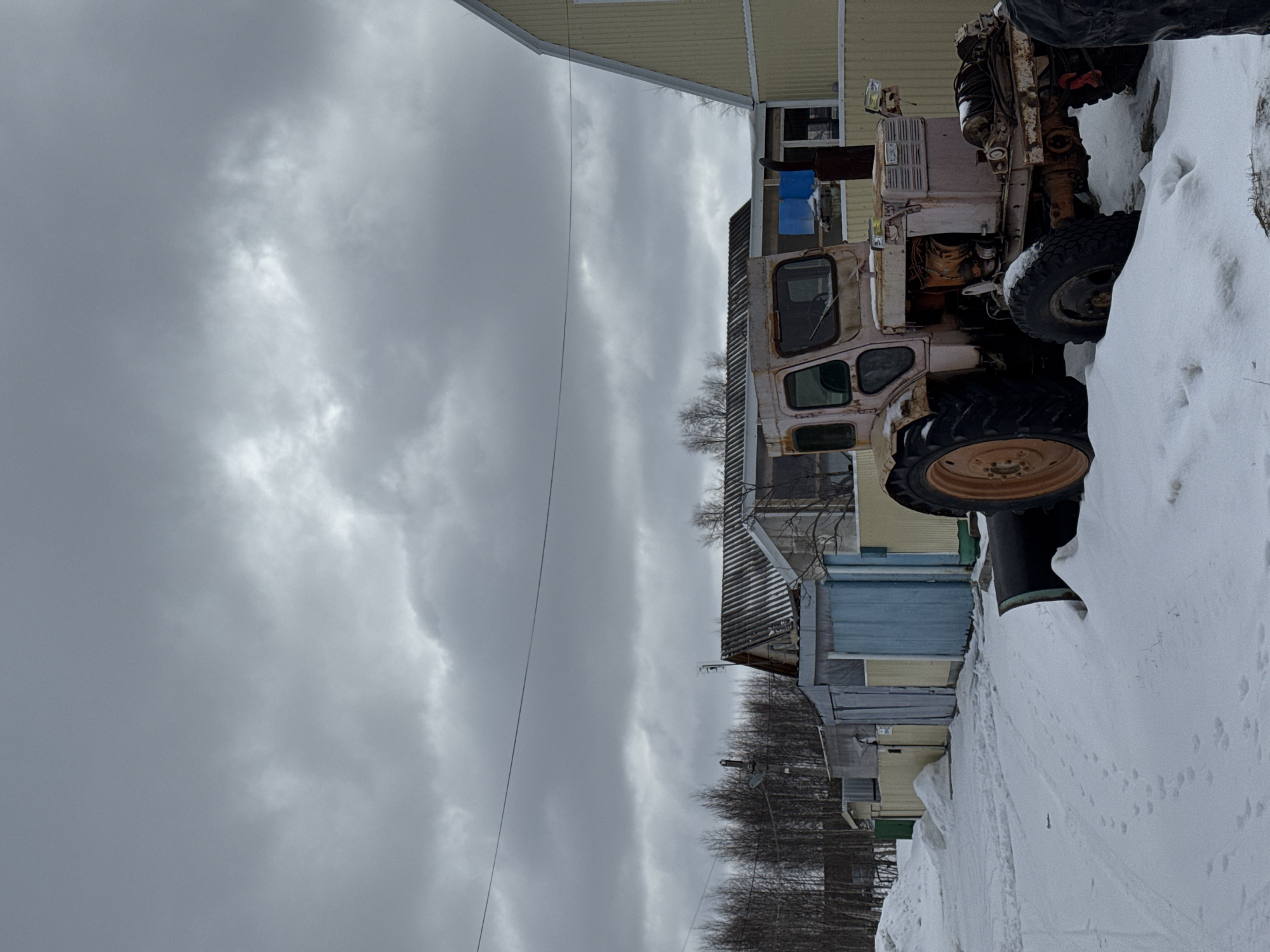 An old, rusty, cream-colored tractor sits partially buried in a snowbank next to a row of weathered, single-story buildings in Vysokiy, Russia. The sky is heavily overcast. Power lines are visible against the cloudy sky. The buildings are of varying colors, primarily light blue, yellow and green. A narrow dirt or gravel path runs alongside the buildings through the snow. Bare trees stand in the background.