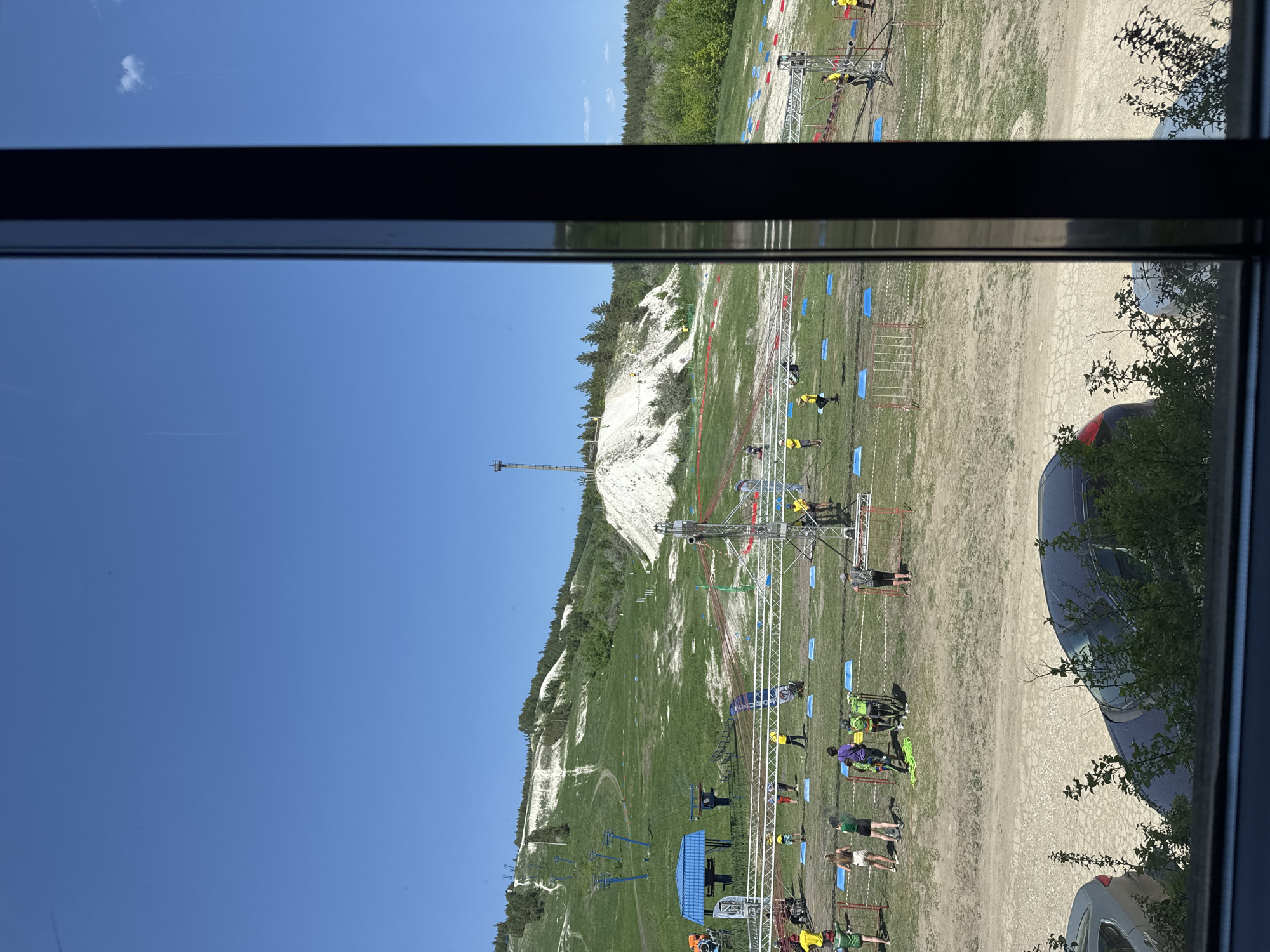 The image shows a view from inside a building, looking out a window. Outside, there is a scene of an obstacle course event on a grassy hill. There are people in various states of action on the course. Structures of scaffolding, blue mats, and red tape are set up as part of the course. A ski lift runs up the hill and a tower is visible on the horizon. Trees are scattered across the landscape. Several parked cars are visible, and some vegetation is obscuring the view. The location is Khvalynsk, Russia.